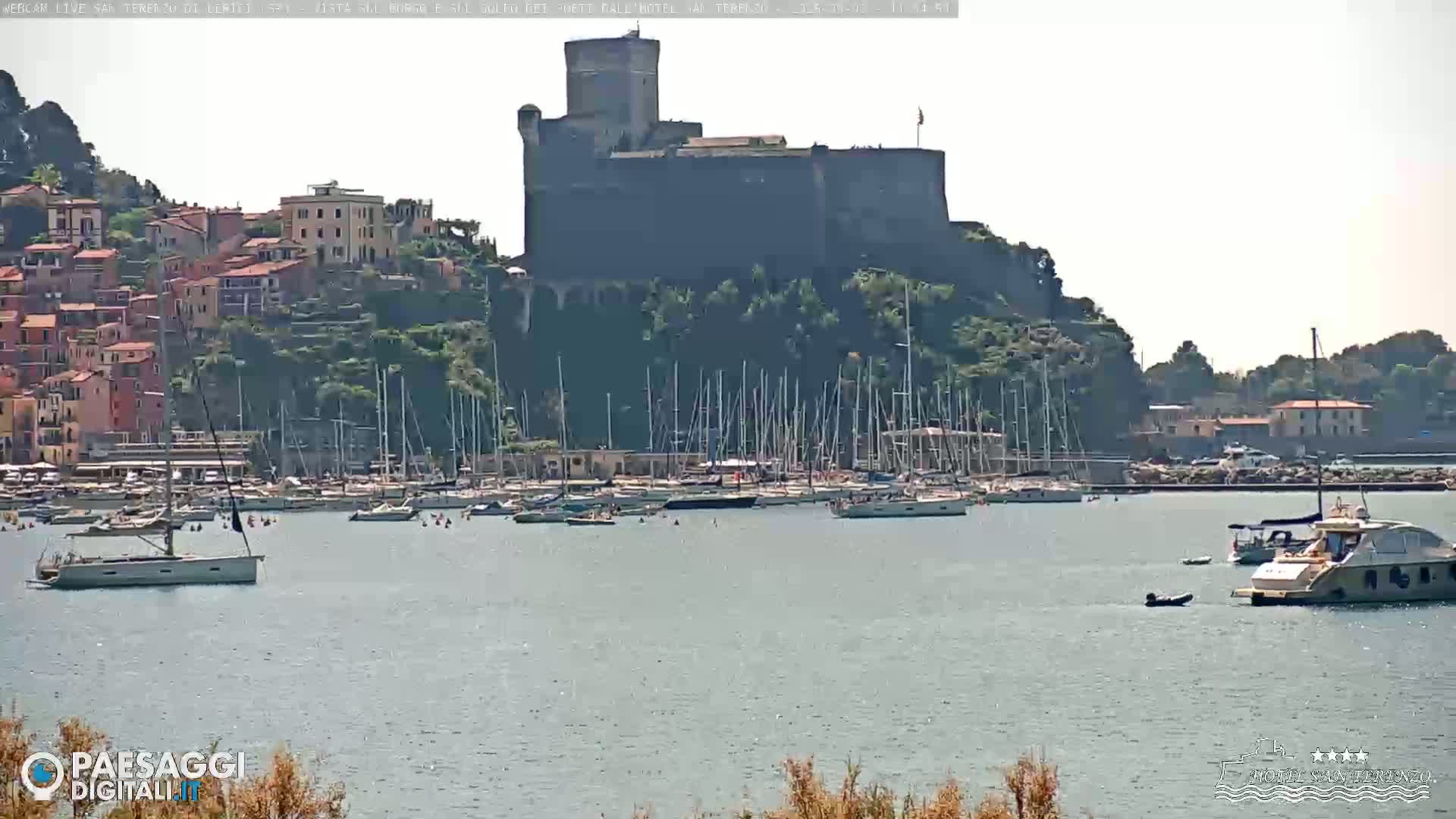 A stone castle sits atop a hill overlooking a calm harbor filled with sailboats and yachts under a clear sky.