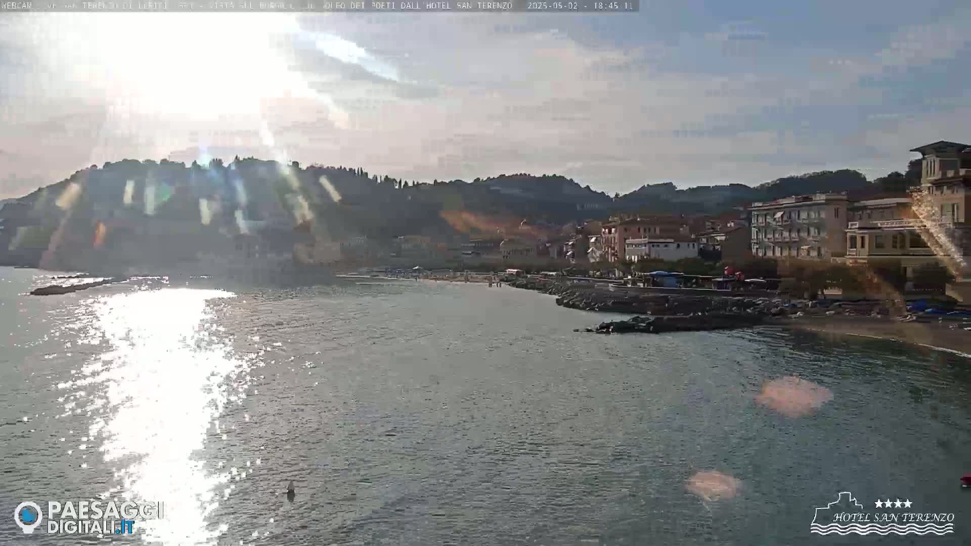 A sunny, partly cloudy day overlooks a calm sea with a beach and buildings nestled against hills in the background.