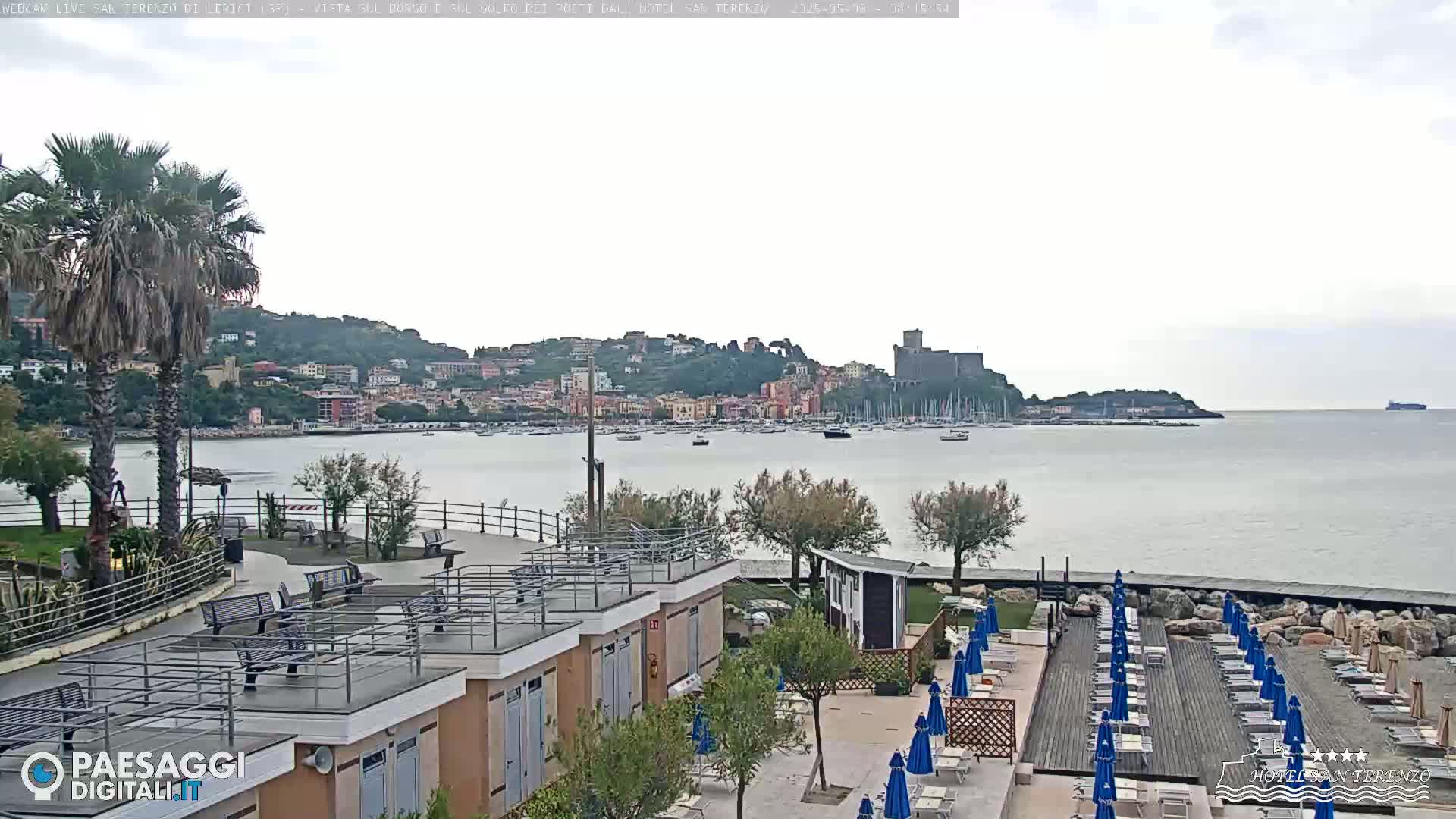 A seaside town with a castle on a hill overlooks a calm bay filled with boats, under an overcast sky; a resort area with lounge chairs and umbrellas is in the foreground.