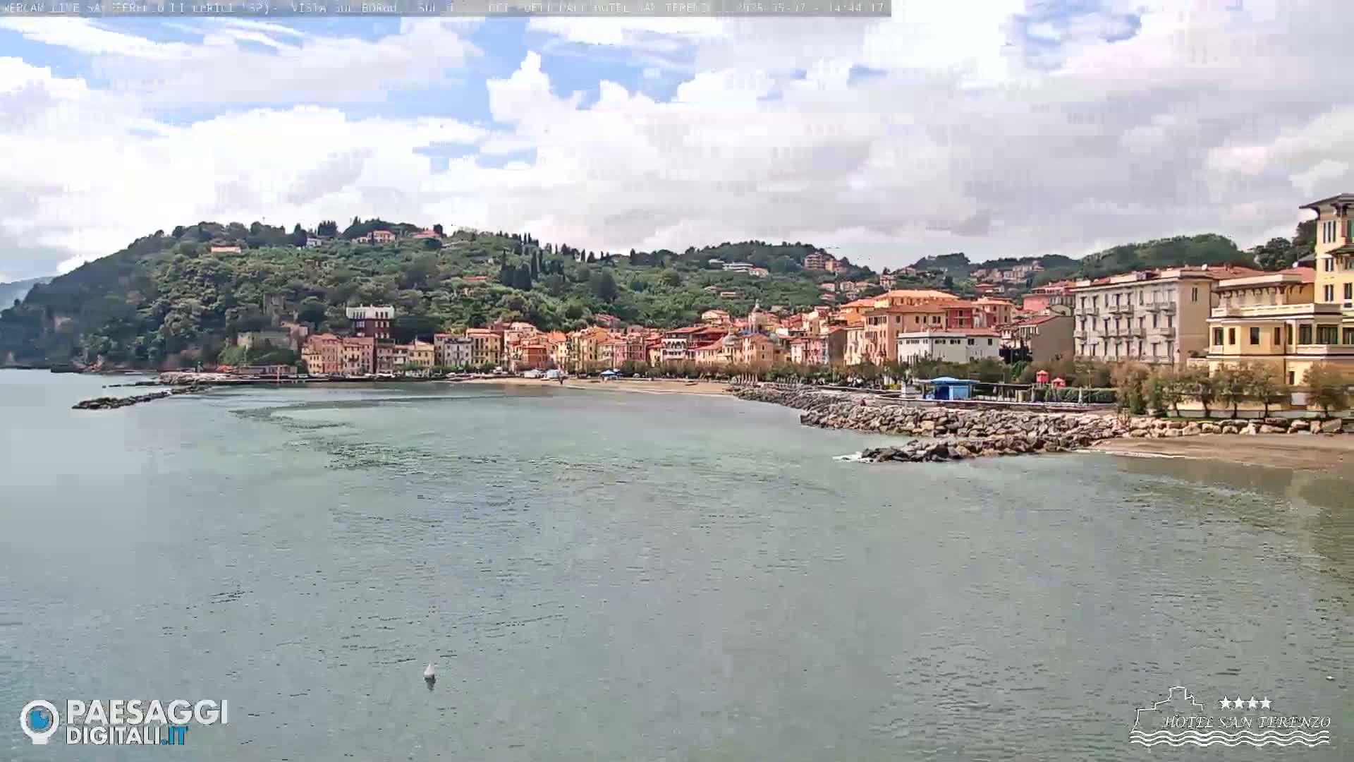 A seaside town with pastel-colored buildings sits on a hillside overlooking a calm bay under a partly cloudy sky.