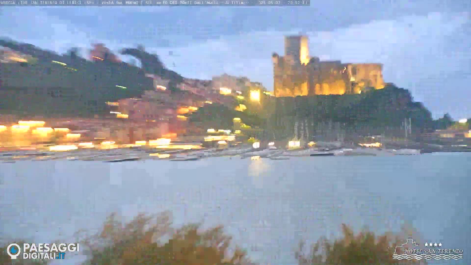 A large stone castle sits atop a hill overlooking a calm body of water with a shoreline of buildings and boats, all illuminated at twilight under an overcast sky.