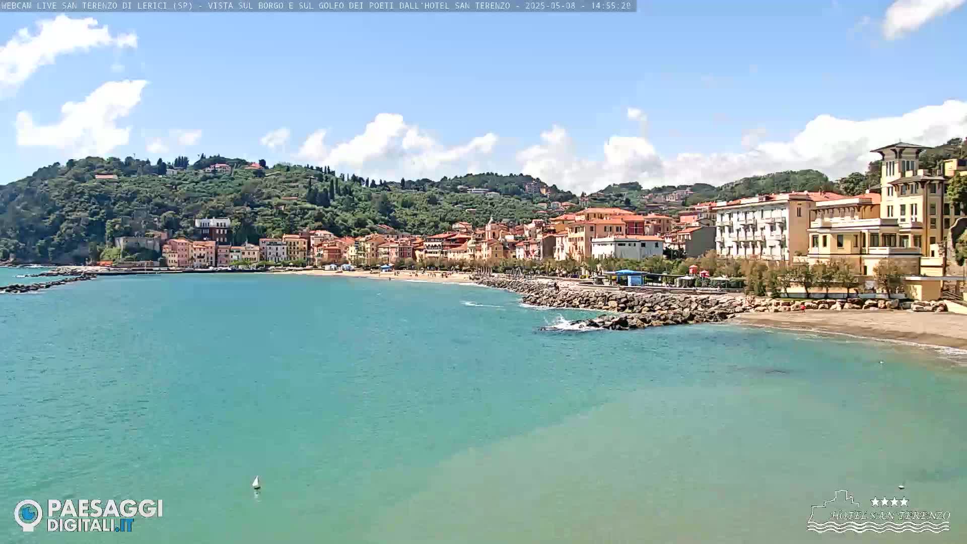 A coastal town with pastel-colored buildings sits on a hillside overlooking a calm, turquoise sea under a partly cloudy sky.