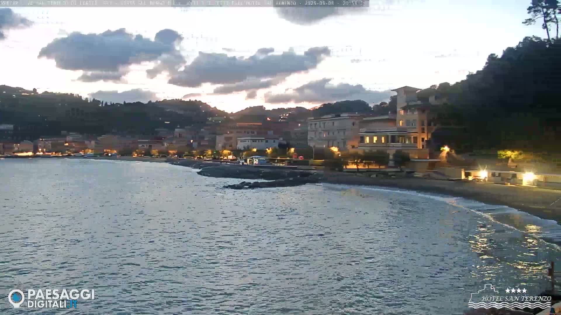 A calm body of water laps a dark-sand beach at dusk, with a village of buildings lit up against a hill under a partly cloudy sky.