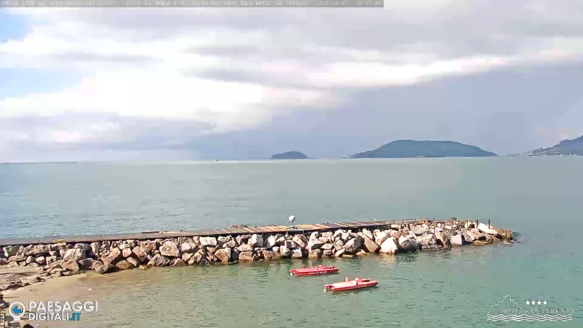 A rocky jetty extends into a calm sea under a partly cloudy sky, with two small red boats moored nearby and distant hills visible on the horizon.