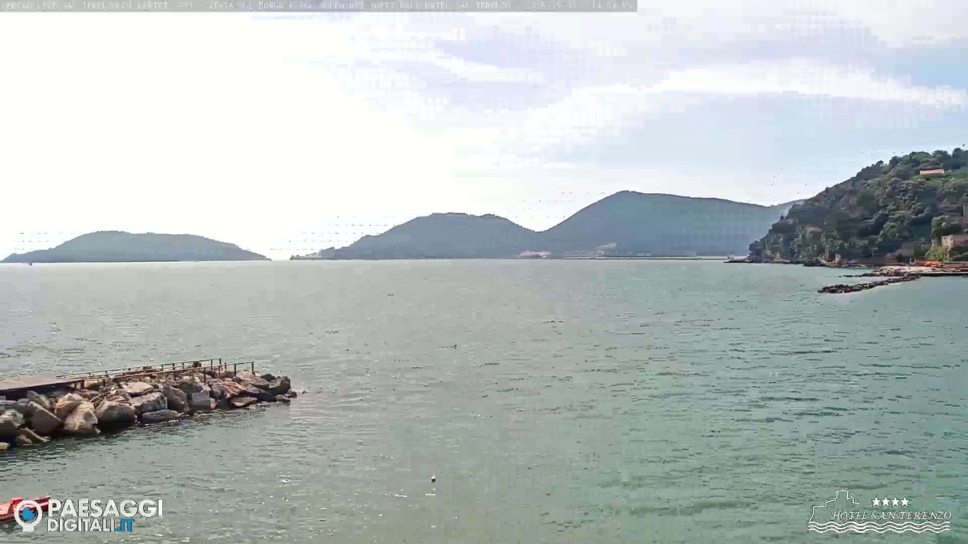 A rocky shoreline extends into a calm, greenish-blue sea, with distant hills and islands under a partly cloudy sky.