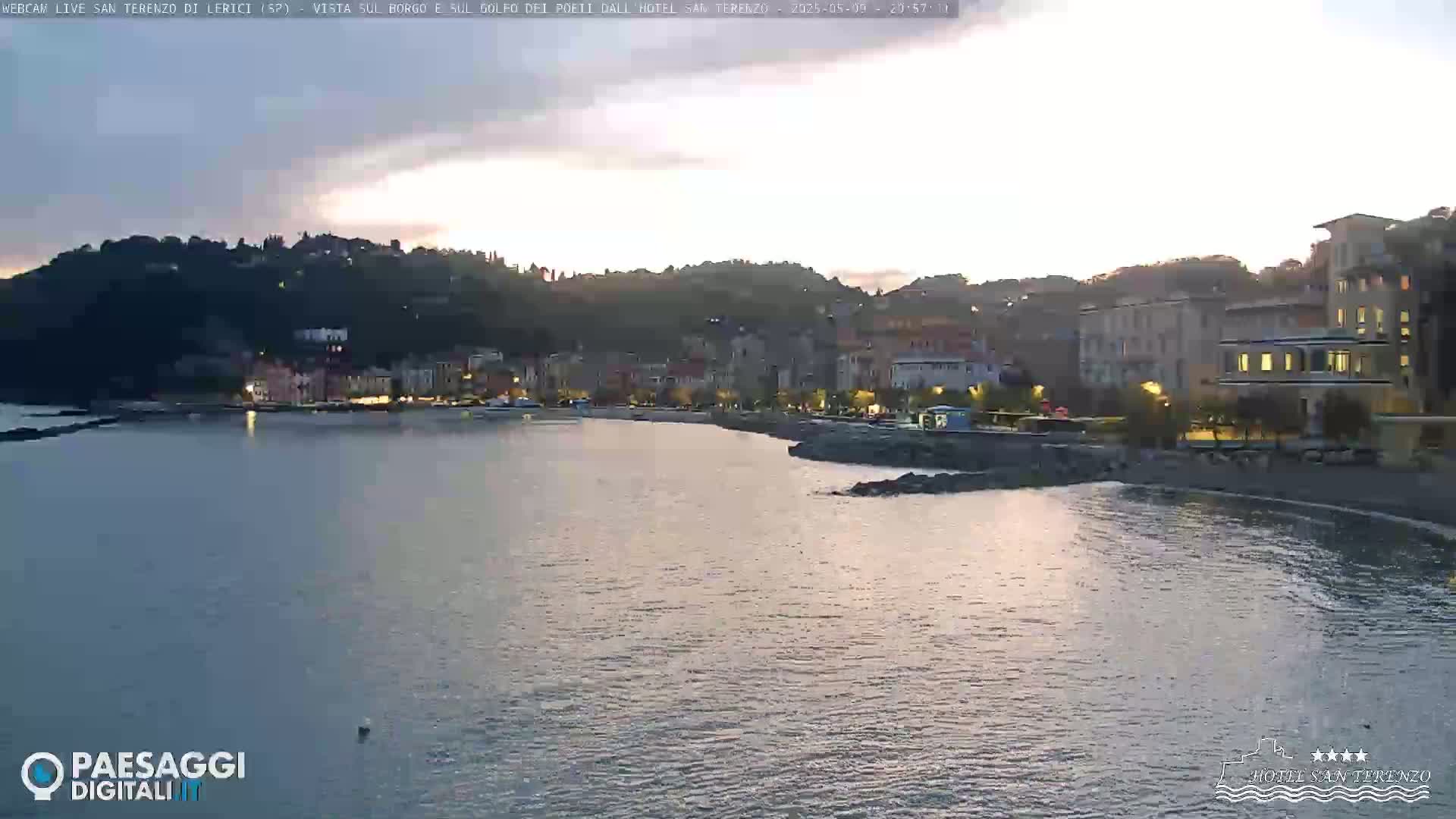 A calm body of water laps a shoreline with a village of buildings nestled against a hillside under a twilight sky.
