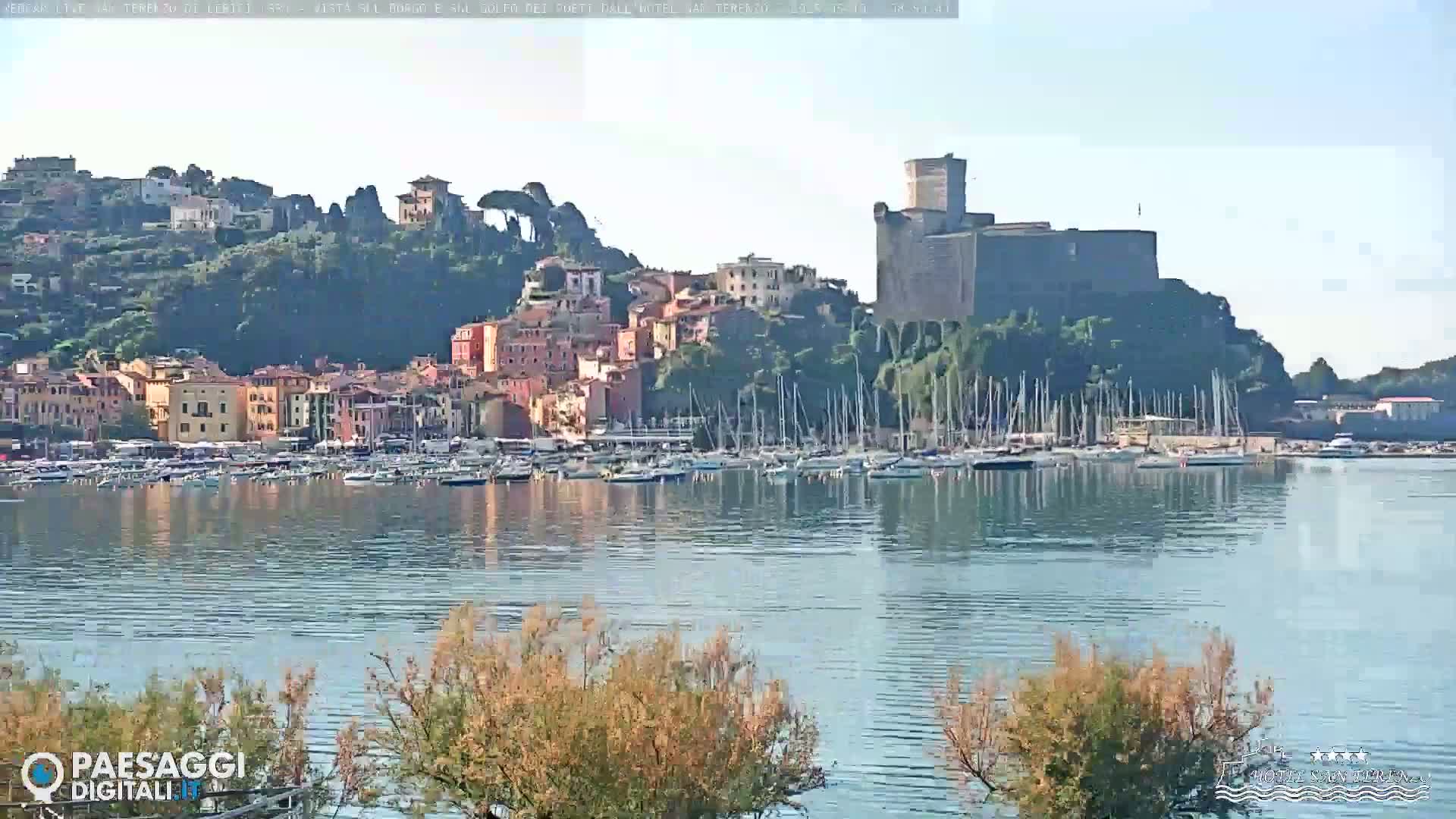 A calm body of water reflects a harbor full of sailboats and a town nestled on a hillside with a castle at its peak on a clear, sunny day.