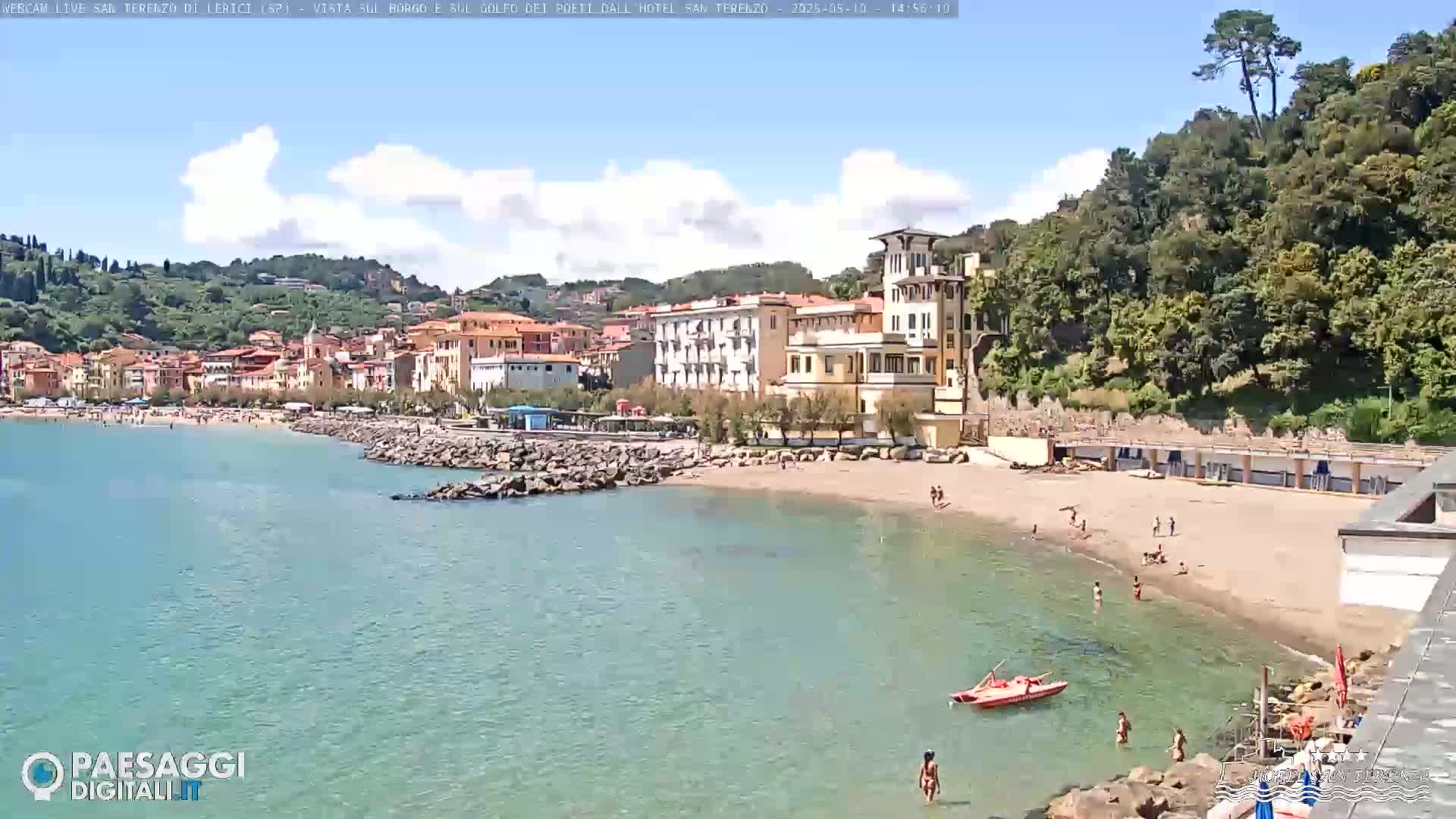 A sandy beach with swimmers in calm, clear water is nestled between a seaside town and a lush green hillside under a partly cloudy sky.