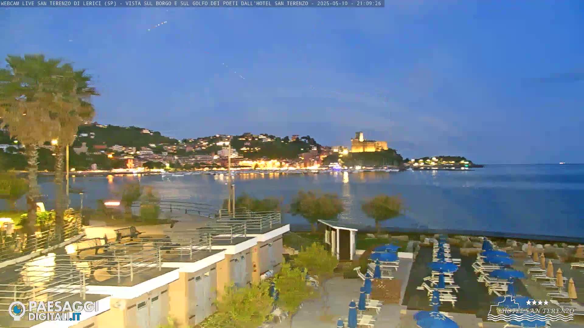 A nighttime view of a calm bay with a town's lights illuminating the shoreline, under a clear twilight sky.