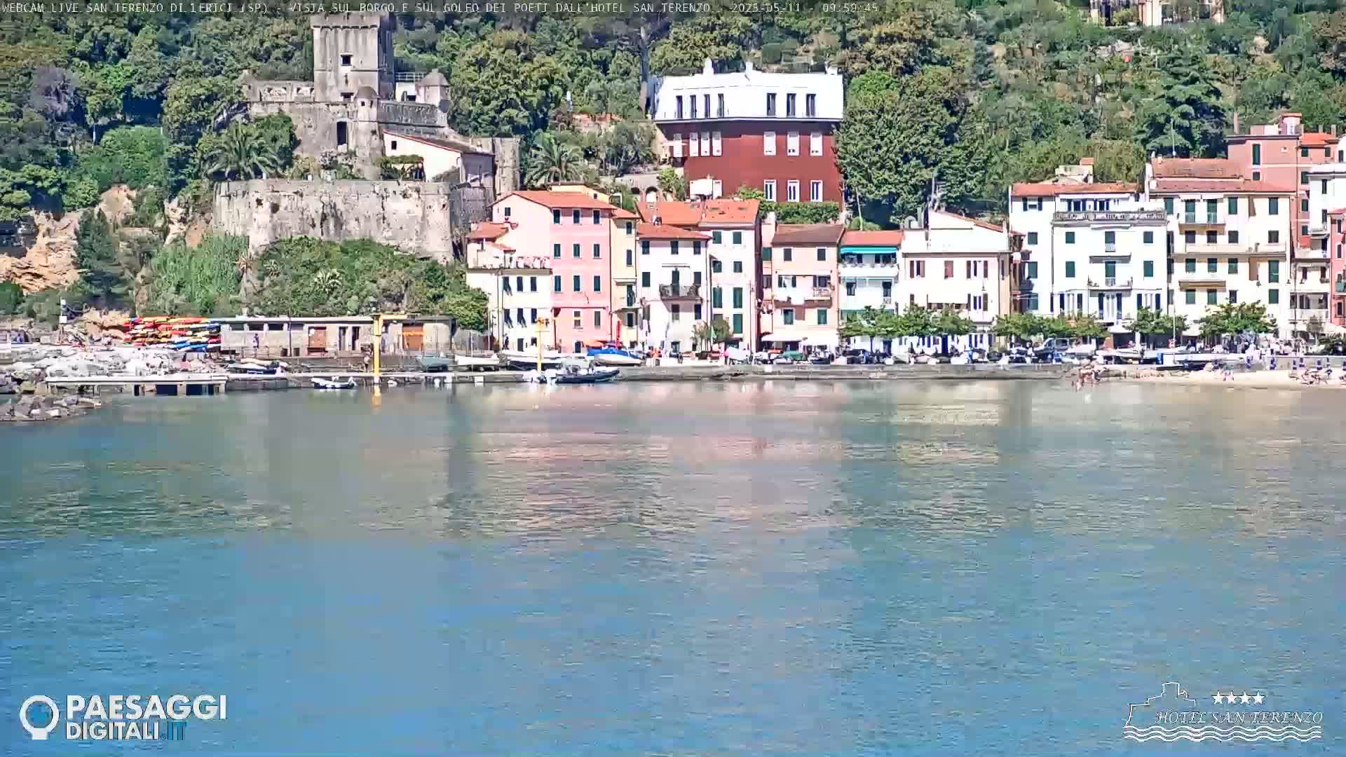 A calm body of water reflects a waterfront town of pastel-colored buildings, a stone castle, and a sandy beach on a sunny day.