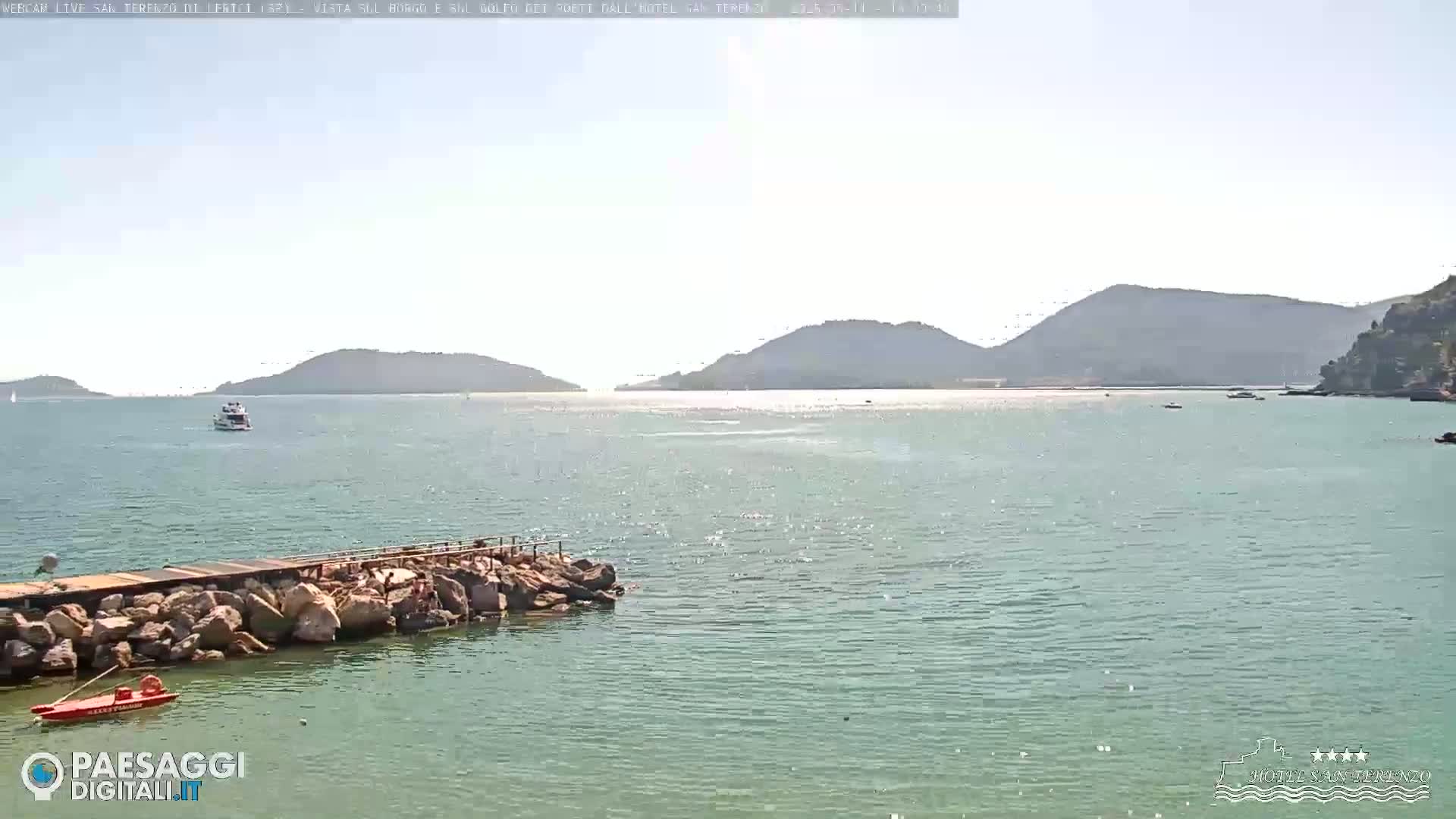 A rocky jetty extends into a calm, light-blue sea, with distant hills and a few boats visible under a bright, sunny sky.
