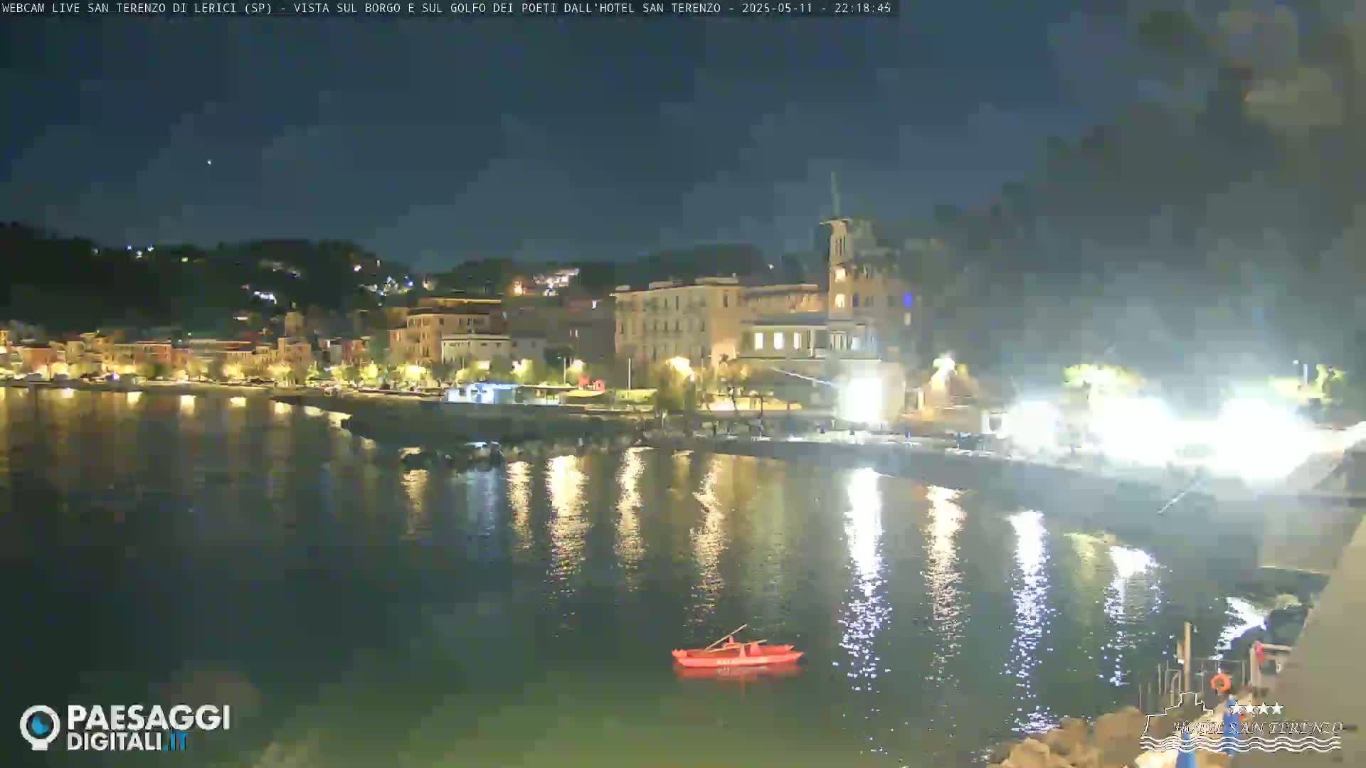 A nighttime view of a calm, illuminated waterfront town with a small red boat on the water.