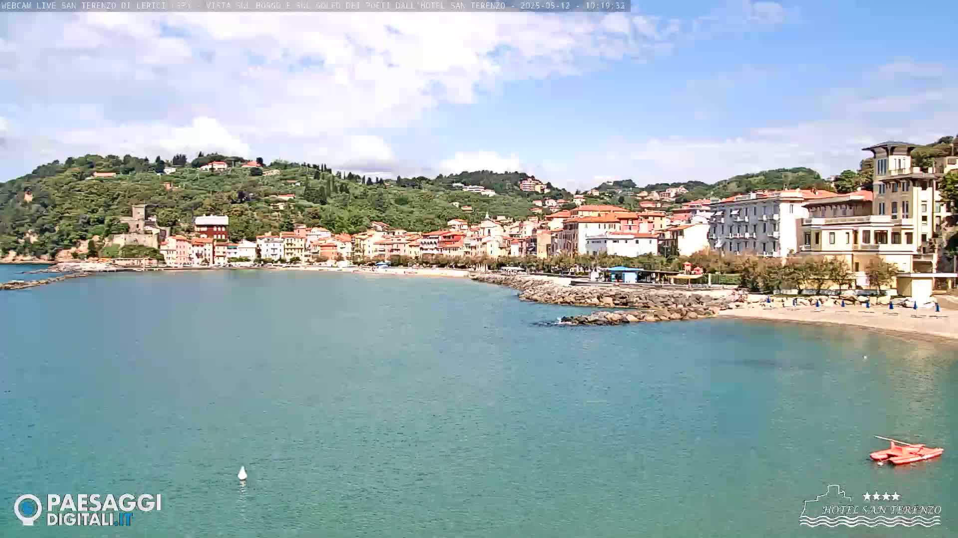 A picturesque coastal town with colorful buildings nestled against a verdant hillside, overlooking a calm, teal sea on a partly sunny day.
