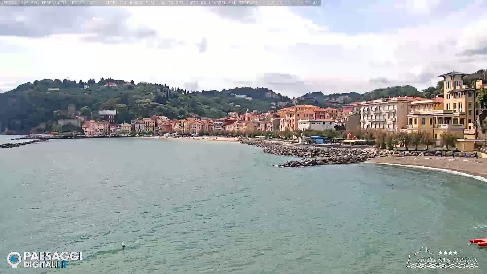 A seaside town with colorful buildings on a hill overlooking a calm bay and a small sandy beach under a partly cloudy sky.