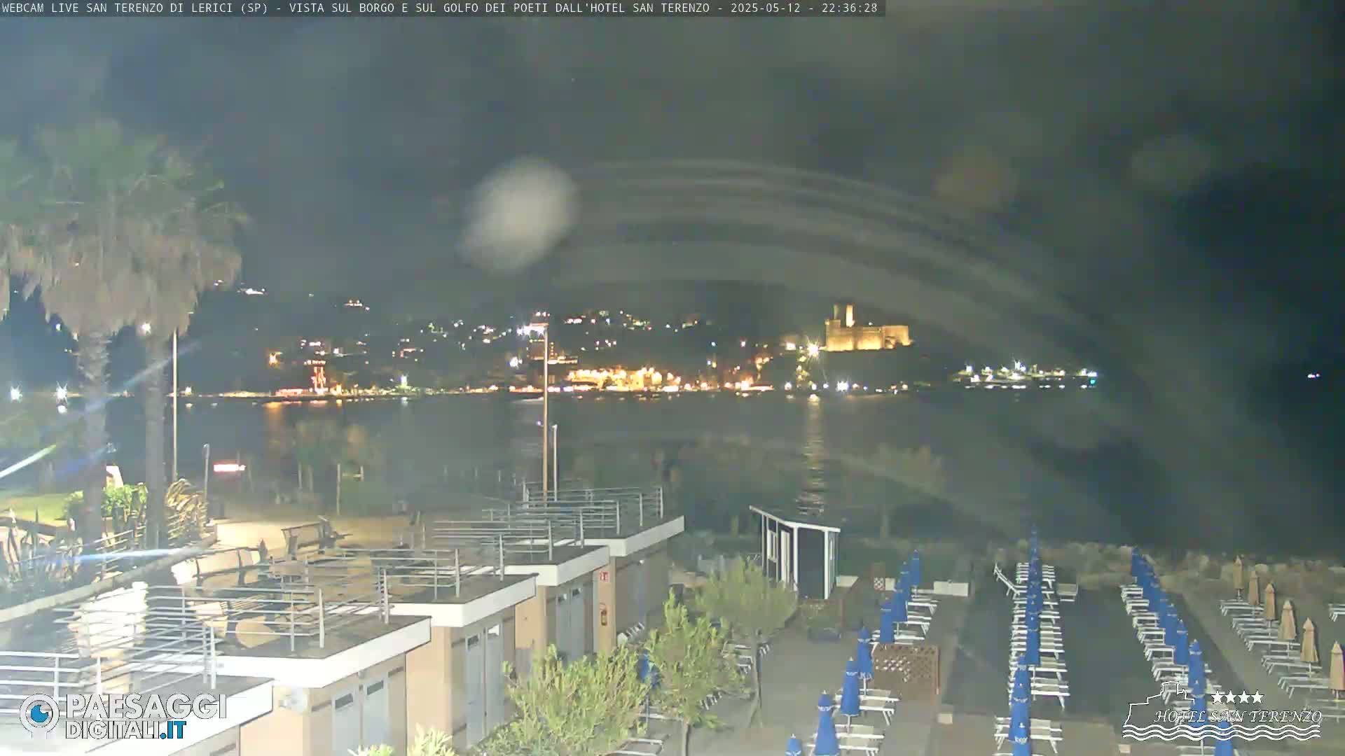 A nighttime seaside view shows a calm body of water, a distant town illuminated with lights, and a beach with rows of closed lounge chairs and umbrellas.