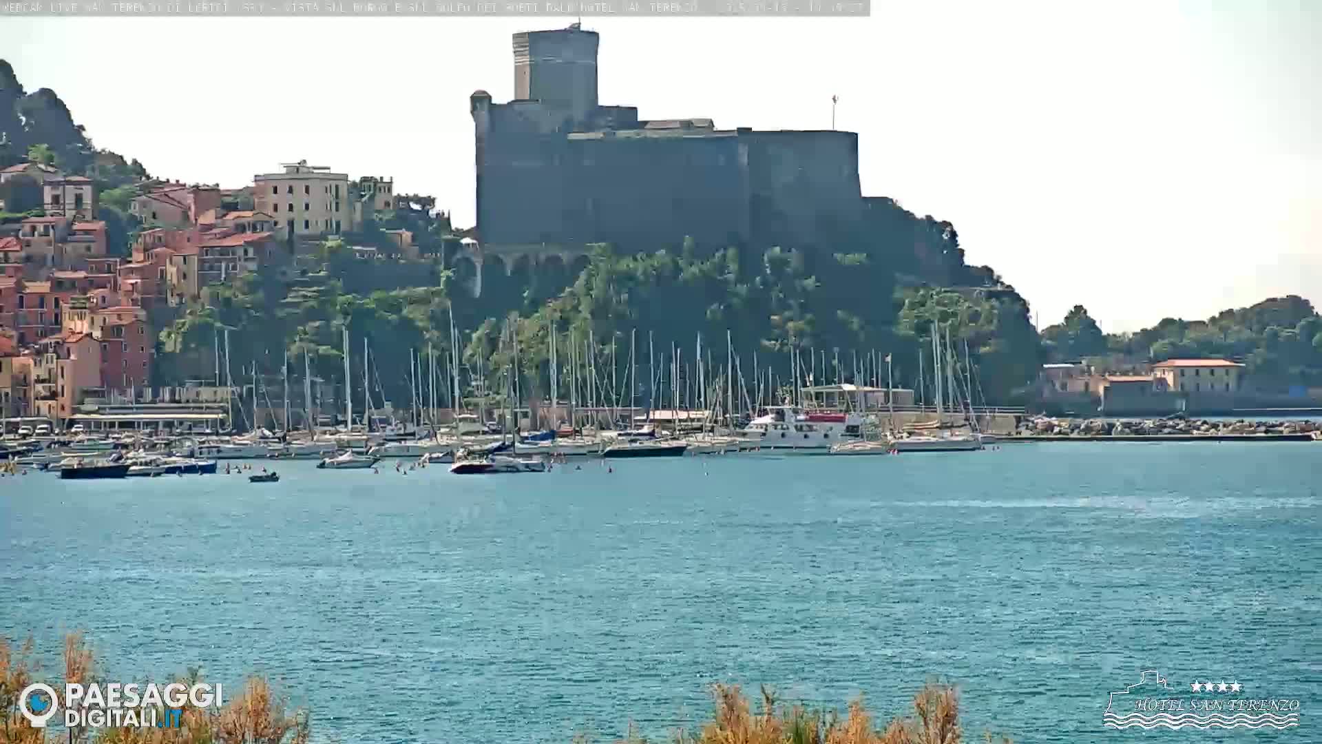 A large stone castle overlooks a harbor filled with sailboats on a sunny day.