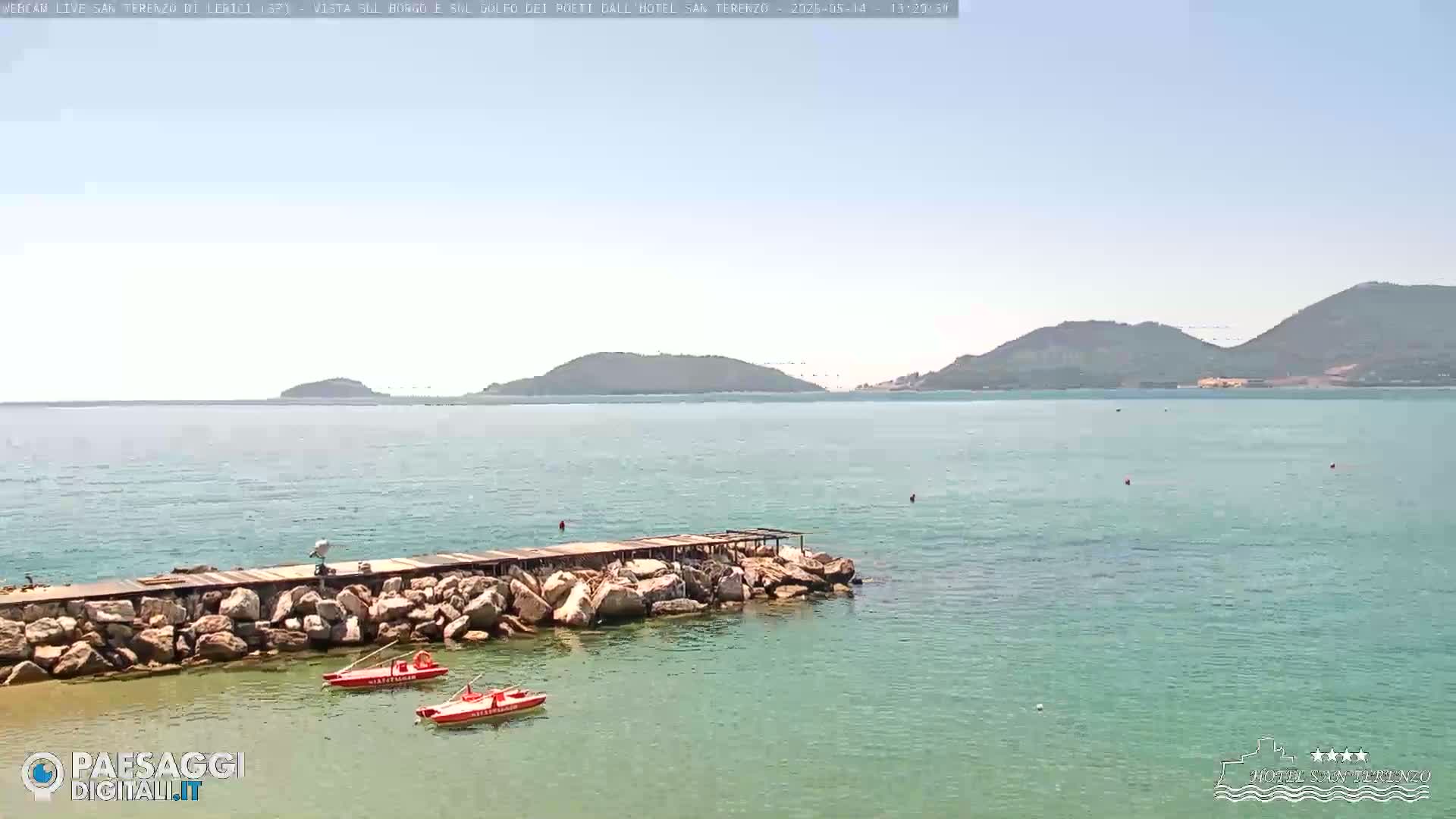 A rocky pier extends into calm, clear turquoise water, with two small red boats moored nearby and distant hills under a bright, sunny sky.