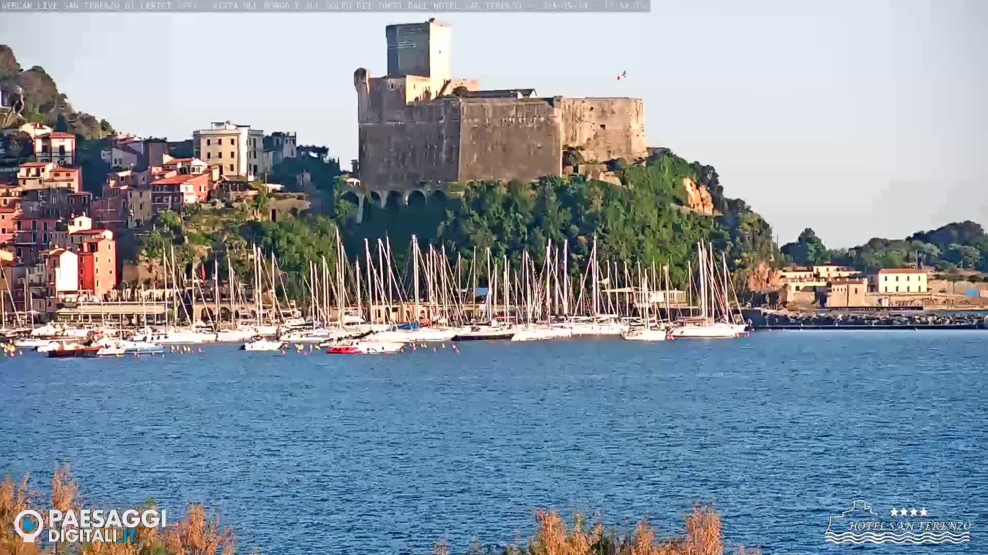 A stone castle sits atop a hill overlooking a harbor filled with sailboats on a calm, sunny day.