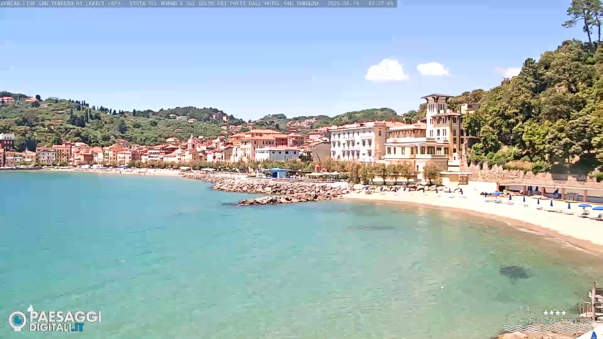 A sandy beach with clear turquoise water laps the shore of a town nestled amongst green hills under a mostly sunny sky.
