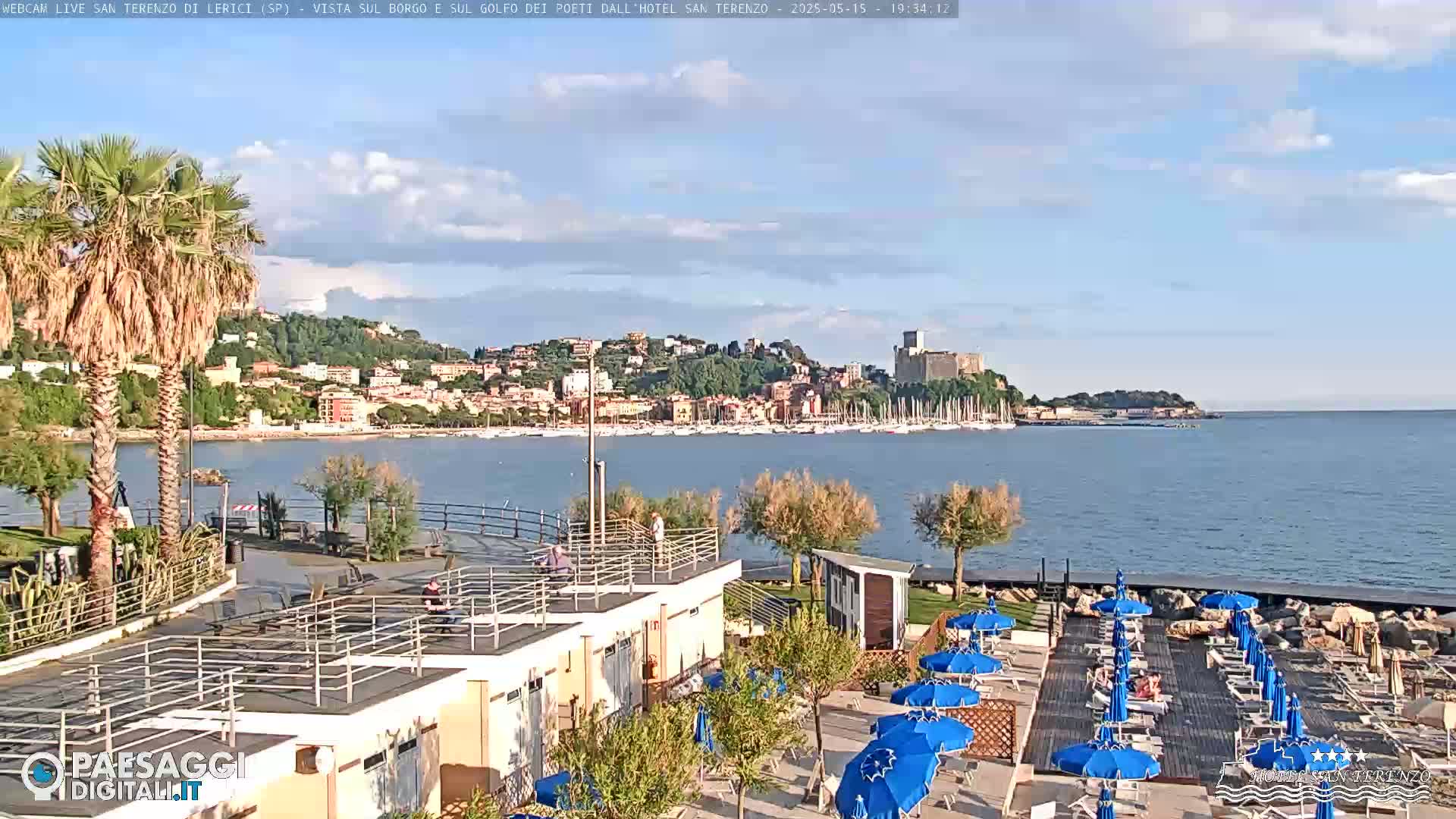 A coastal town with a harbor full of boats is visible across a calm body of water, under a partly cloudy sky, from a beachside vantage point with several blue umbrellas and lounge chairs.