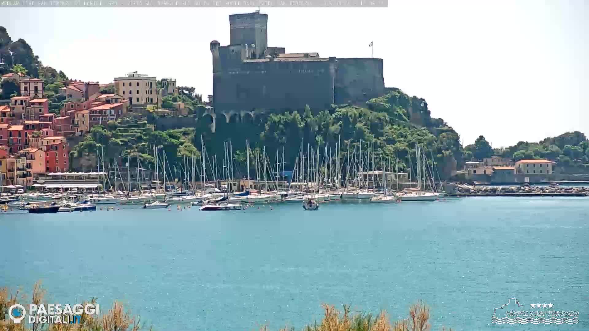 A stone castle sits atop a hill overlooking a harbor filled with sailboats on a sunny day.
