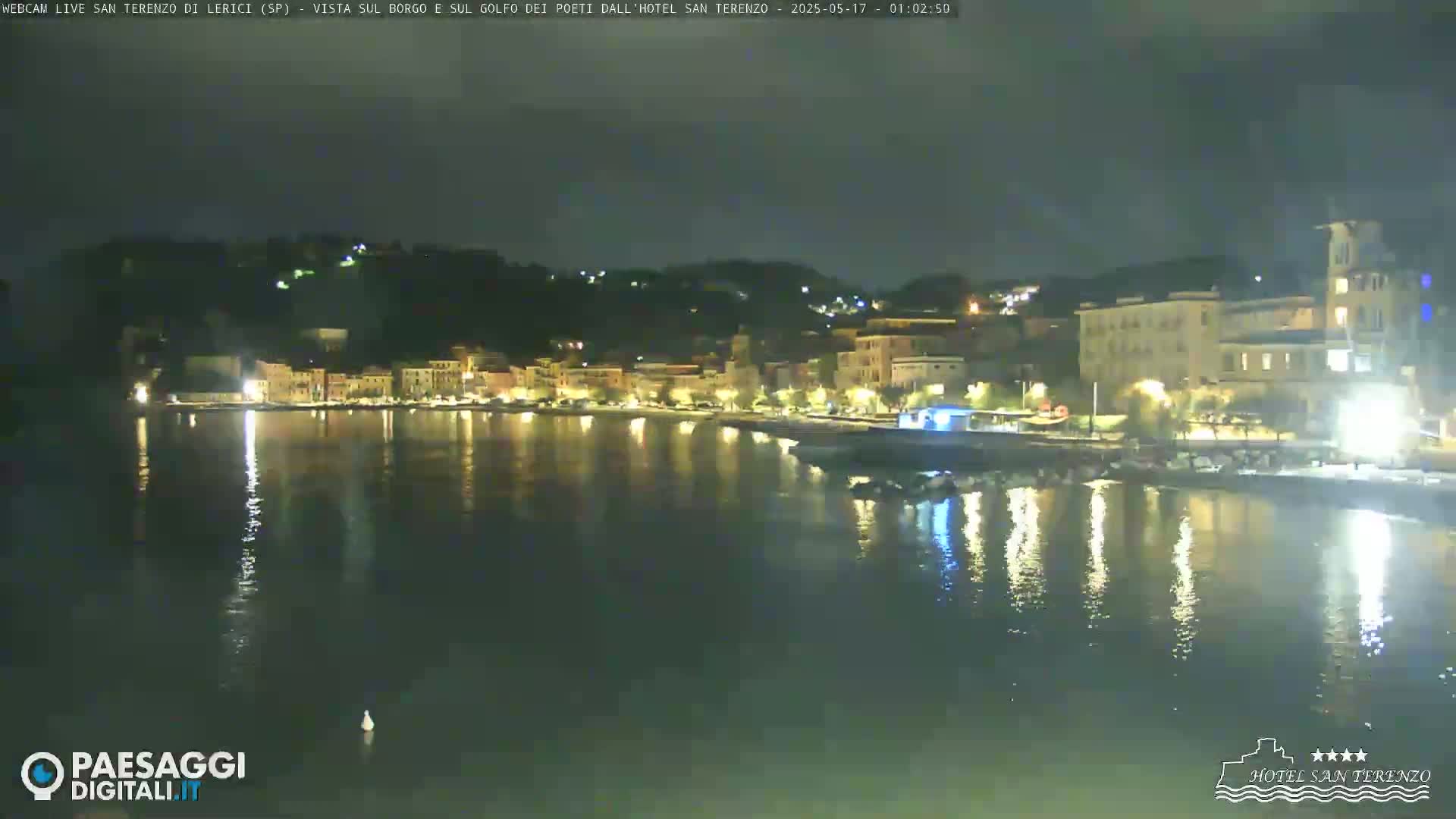 A nighttime view of a waterfront town with buildings illuminated along the shore and their lights reflected in the calm water.