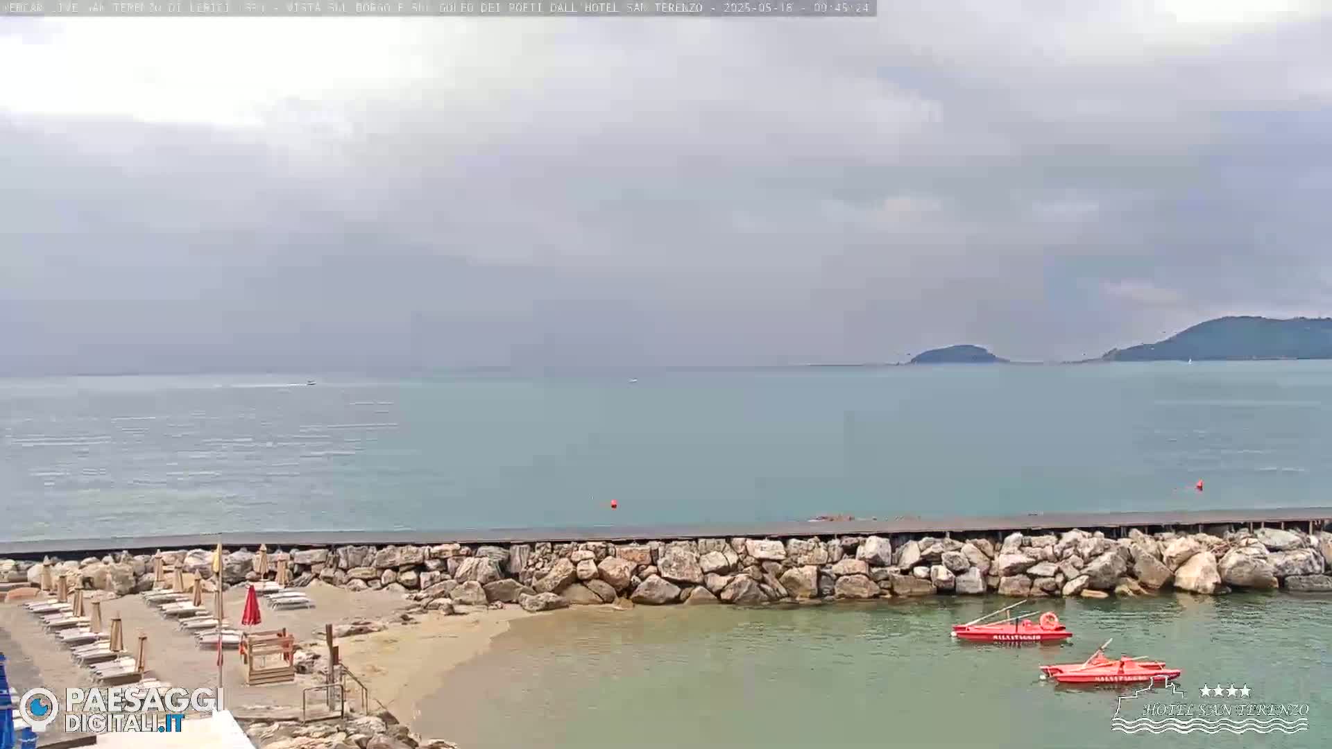A rocky seawall protects a small, sandy beach with lounge chairs and umbrellas under a cloudy sky, overlooking calm, teal water with distant hills.