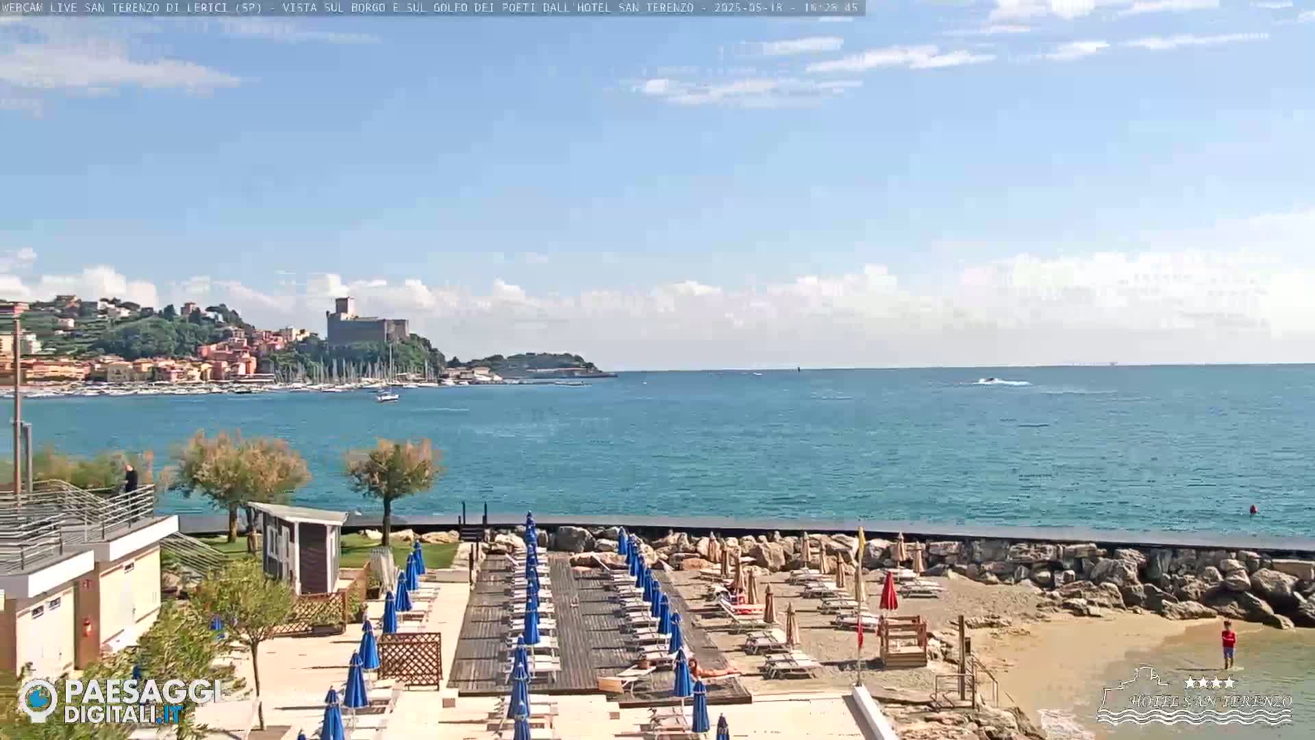 A beach with blue umbrellas and lounge chairs overlooks a calm, blue sea with a town and castle visible in the distance under a partly cloudy sky.