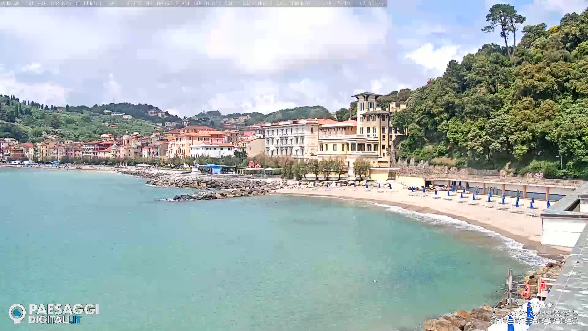 A sandy beach with turquoise water, lined with beach umbrellas, and buildings on a hillside under partly cloudy skies.