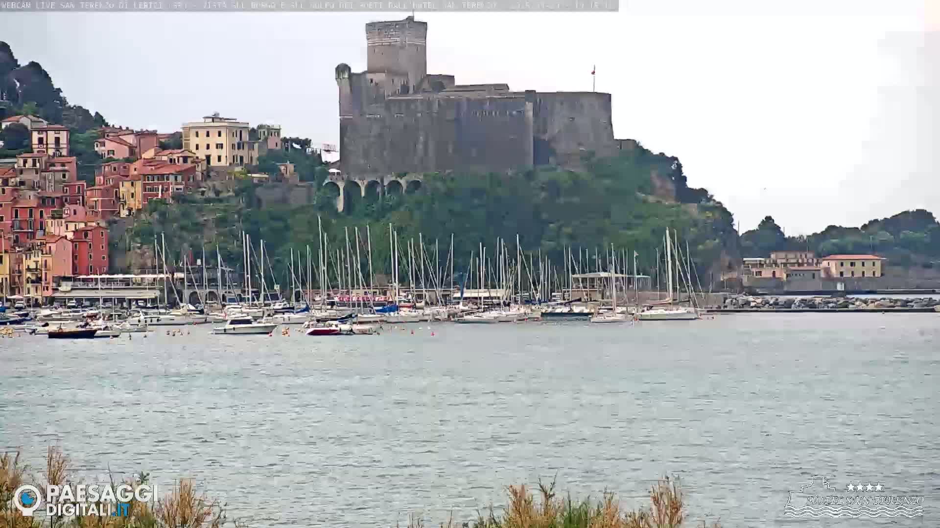 A stone castle overlooks a harbor filled with sailboats on a calm, overcast day.
