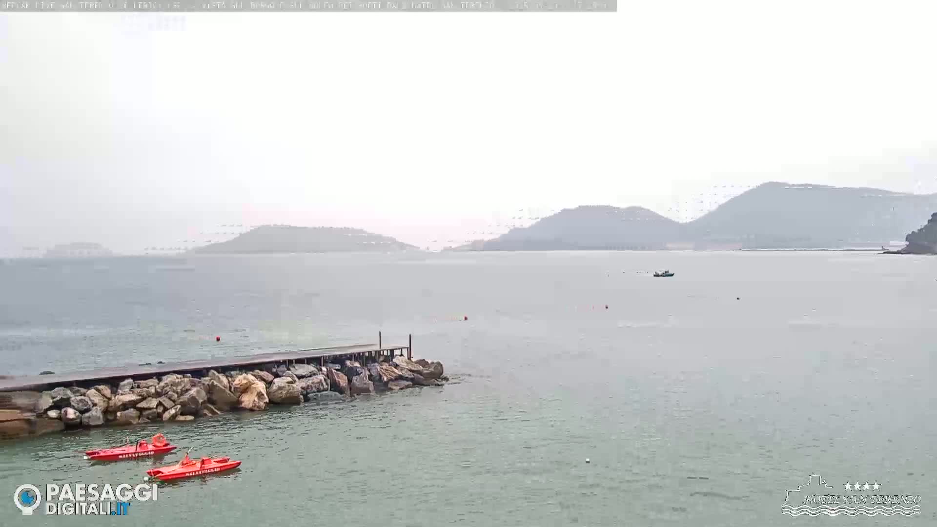 Two red rescue boats are moored near a short pier jutting into a calm, overcast sea, with distant hills visible in the hazy background.
