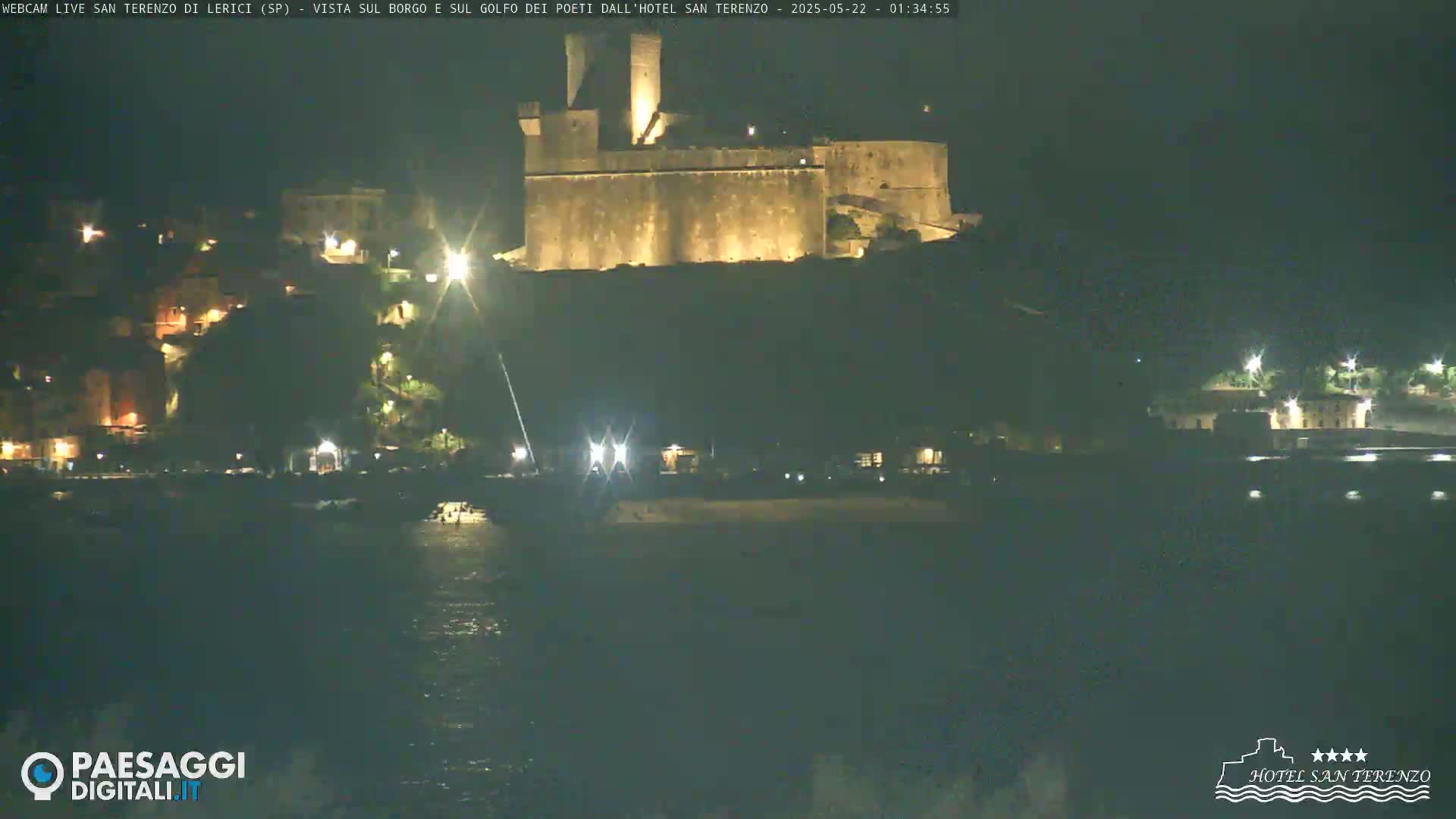 A nighttime view of a coastal town with a large, illuminated fortress overlooking a dark body of water.