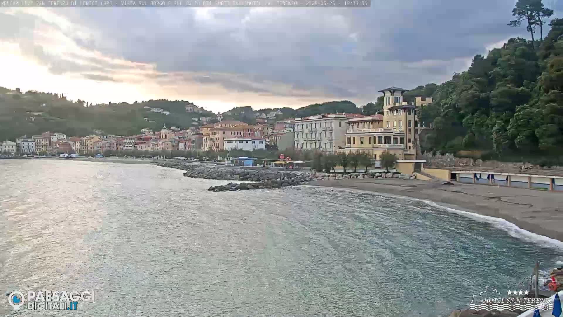 A seaside town nestled against a hillside under a partly cloudy sky, with calm water lapping a sandy beach.