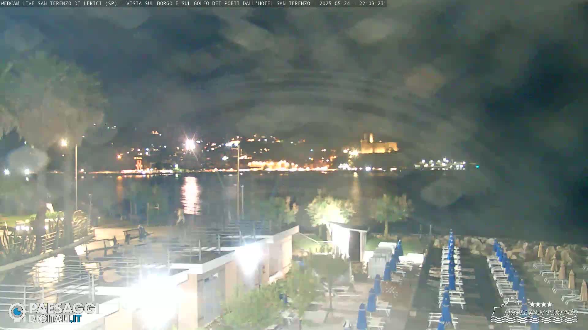 A nighttime view of a beach with rows of lounge chairs and umbrellas, overlooking a calm body of water and a brightly lit town across the water under a partly cloudy sky.