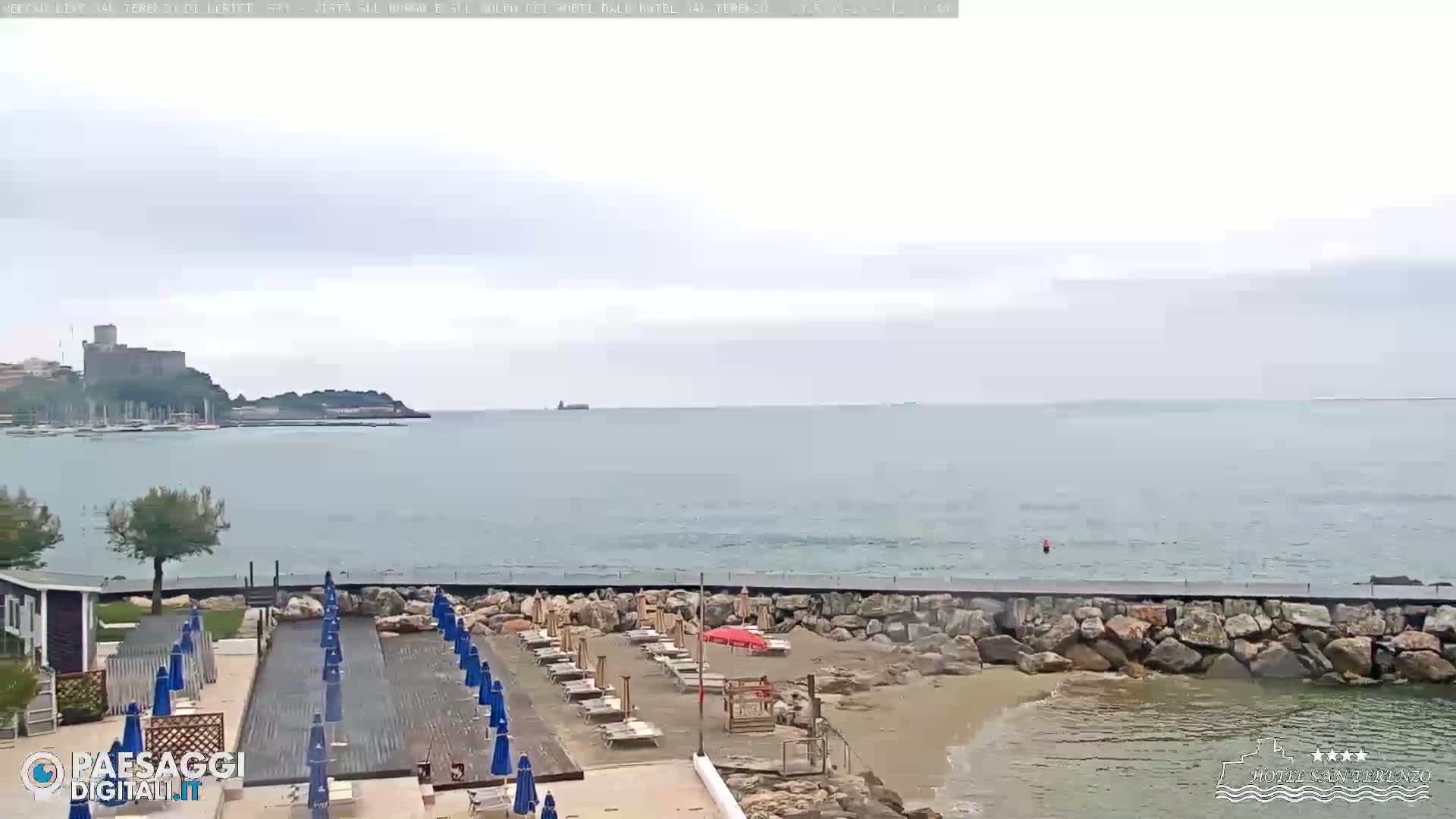 A mostly cloudy day shows a beach with rows of closed umbrellas and lounge chairs next to a rocky seawall overlooking calm ocean water and a distant castle.