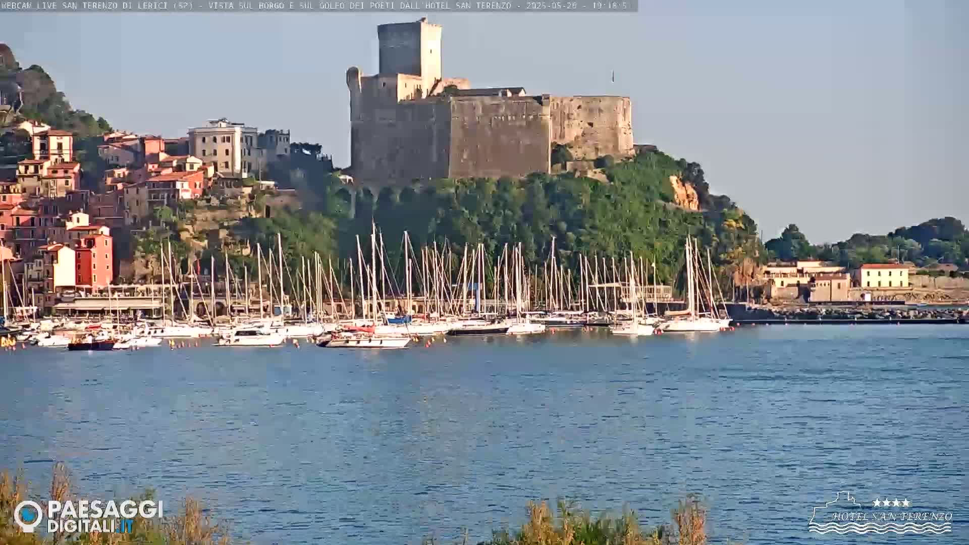 A stone castle sits atop a hill overlooking a harbor filled with sailboats on a calm, sunny day.