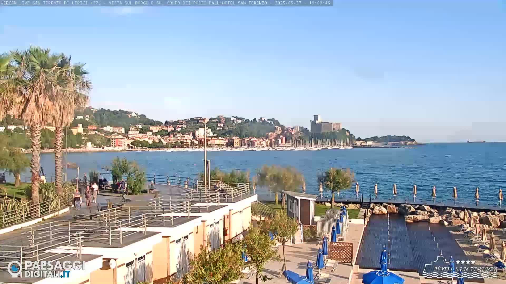 A seaside resort with a swimming pool area overlooks a calm bay filled with boats, a town nestled on a hillside, and a castle in the distance under a clear blue sky.