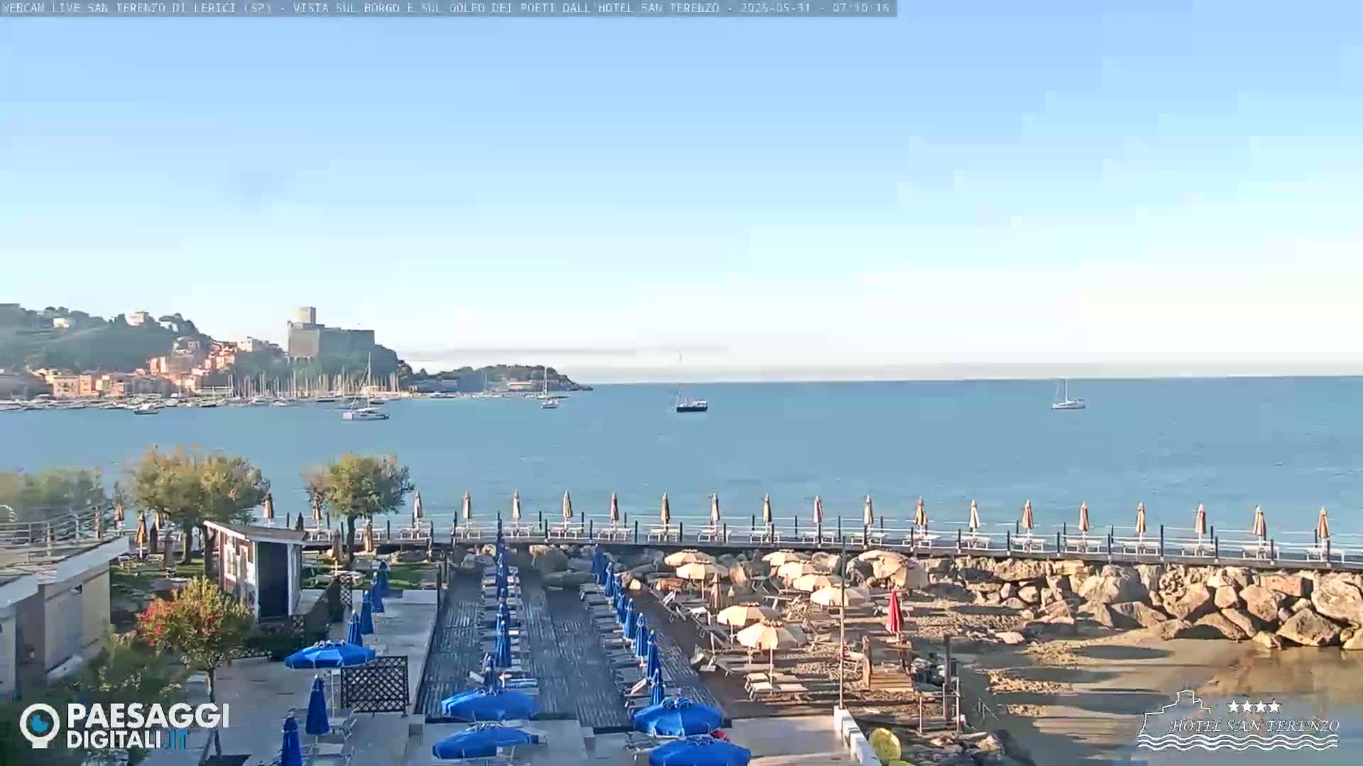 A seaside resort with blue umbrellas on a sandy beach overlooks a calm bay with a town and a castle on a distant hill under a clear sky.
