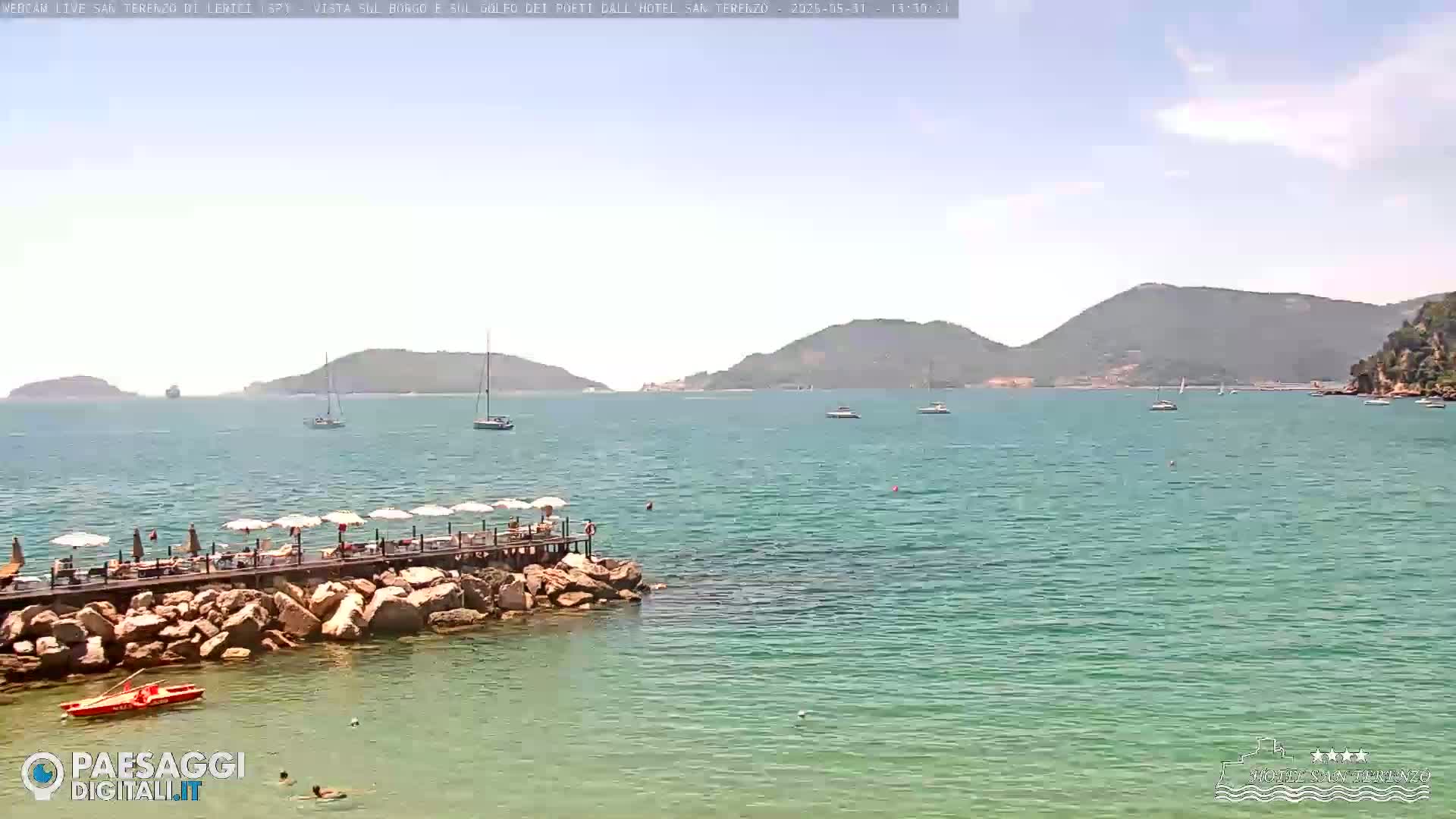 A clear, sunny day shows a calm, turquoise sea with several sailboats and a rocky shoreline featuring a pier with umbrellas and people.