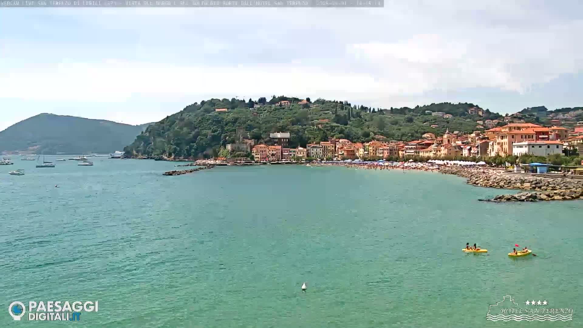 A coastal town with a sandy beach is visible across a calm, teal-colored body of water under a partly cloudy sky.