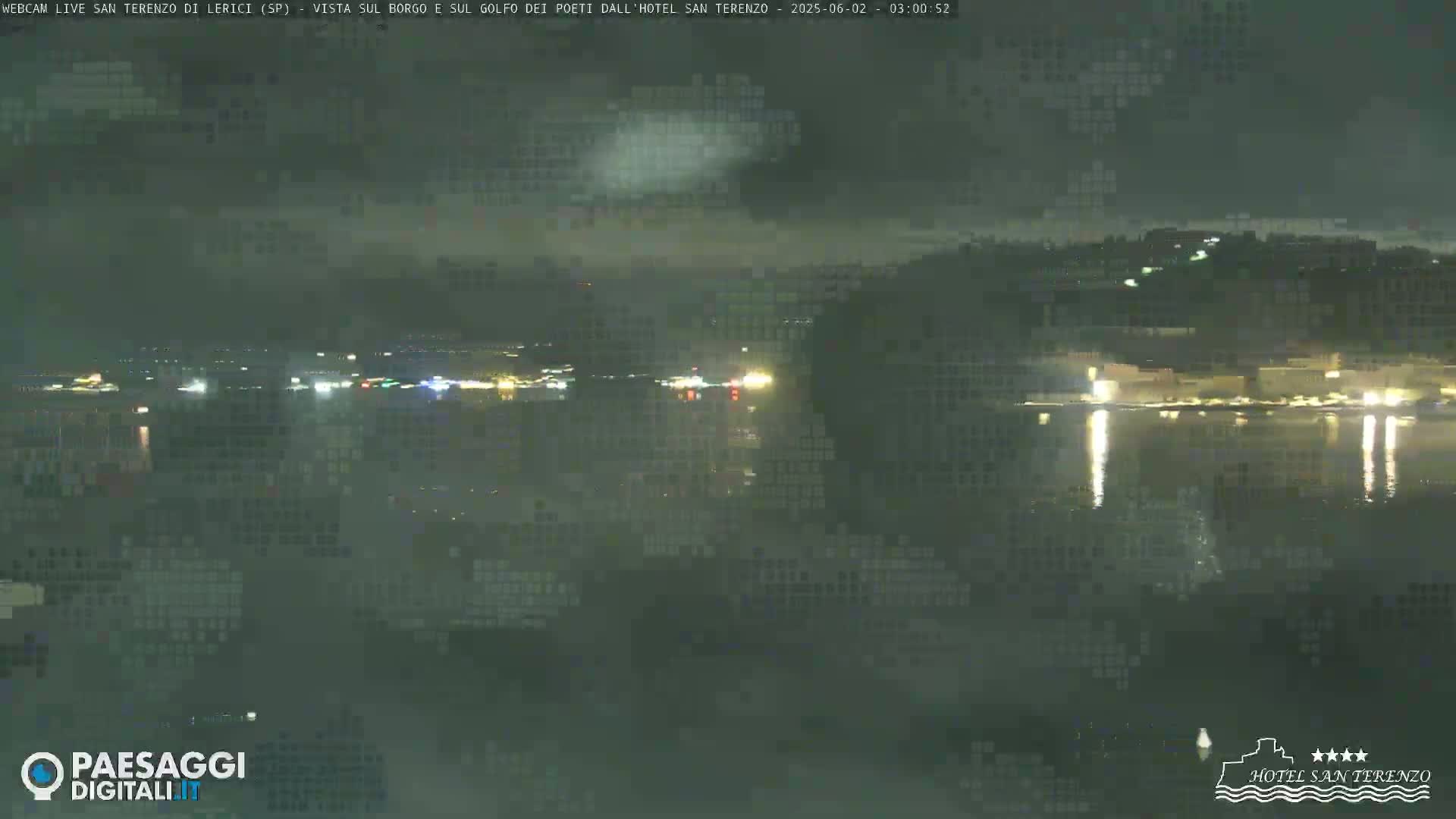 A nighttime view of a calm body of water reflecting the lights of a town on the opposite shore, under a hazy sky.