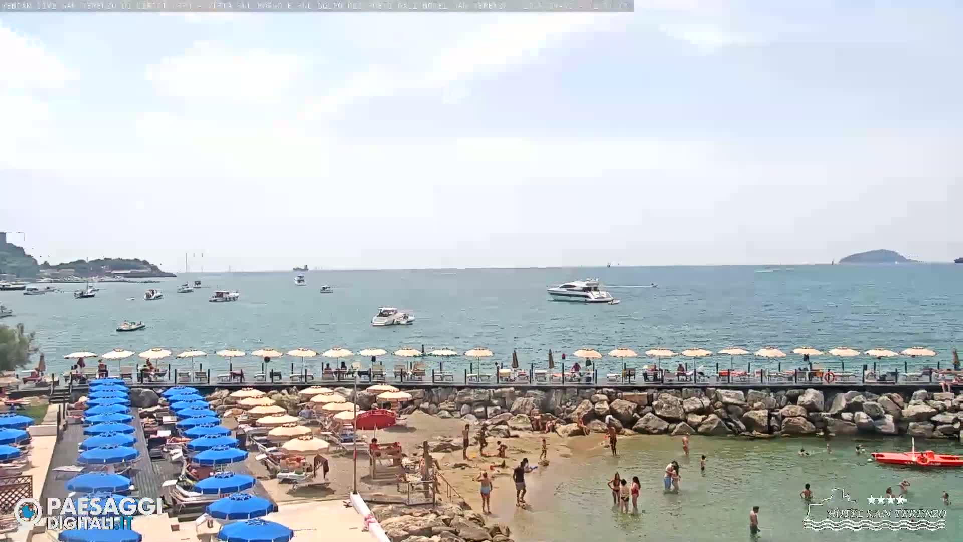 A beach with numerous umbrellas and sunbathers is situated beside calm blue water with boats and small islands in the distance under a mostly clear sky.