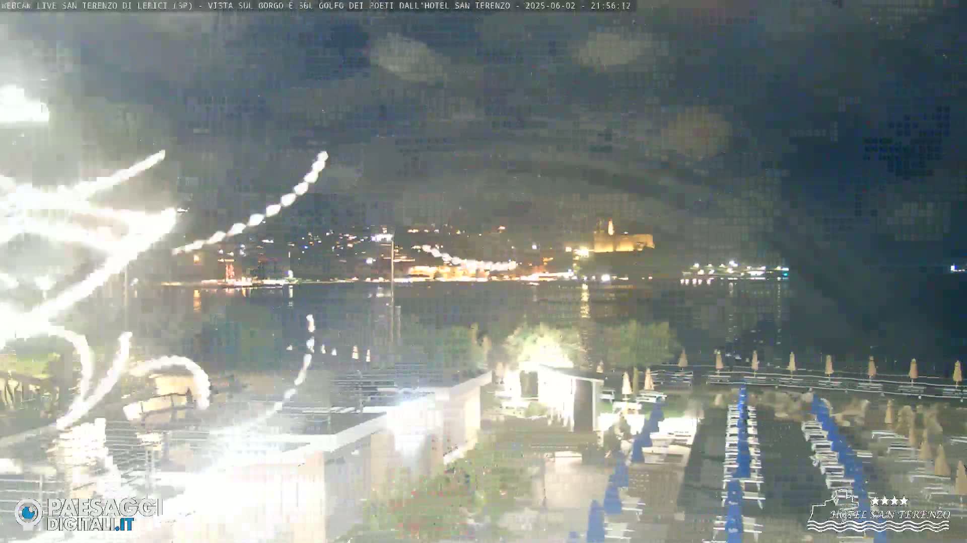 A nighttime view of fireworks over a calm body of water with a town illuminated in the background, and beach chairs in the foreground.