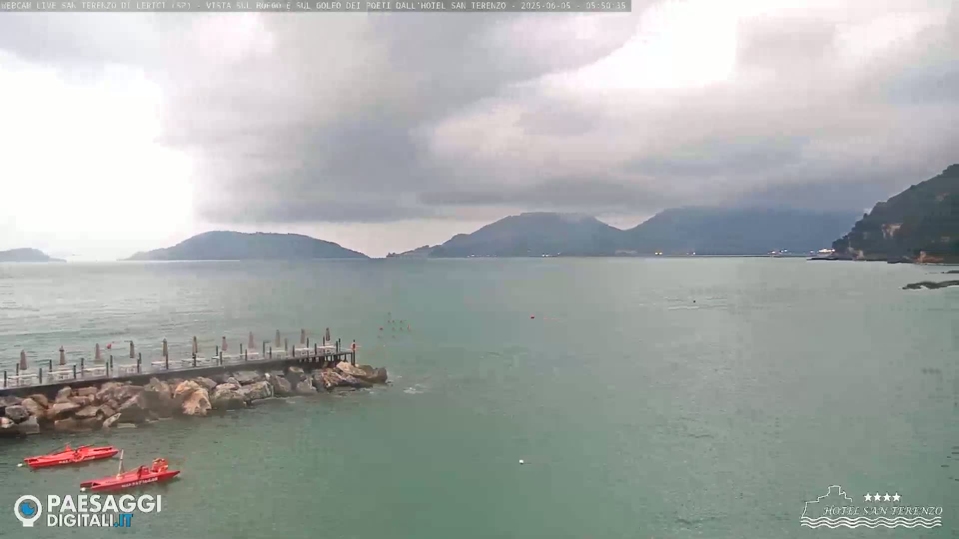 A calm, overcast day shows a body of water with a long dock extending from a rocky shore toward distant mountains.