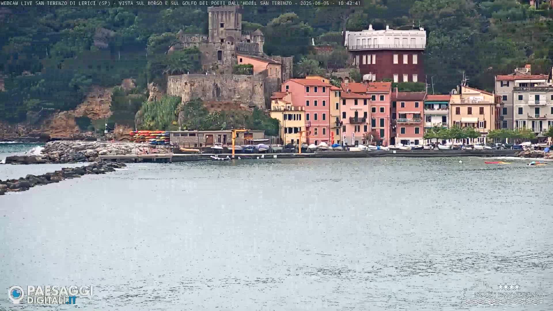 A waterfront town with colorful buildings, a stone jetty, and several boats is seen from across a calm body of water on an overcast day.