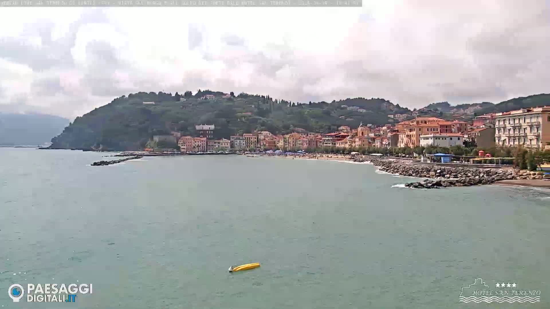 A coastal town nestled against a verdant hillside, with a calm sea and overcast skies, features a sandy beach and a small yellow boat floating offshore.