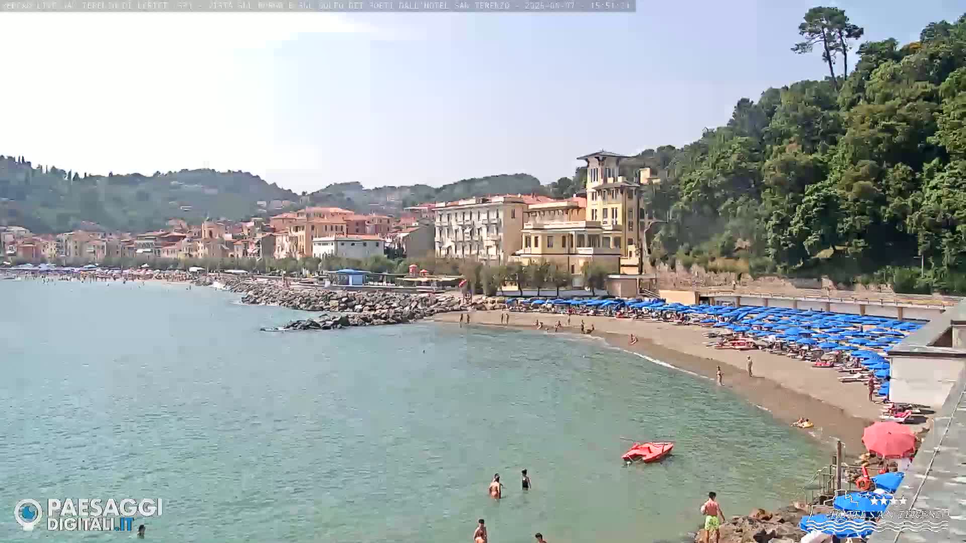 A sunny beach scene shows a sandy shoreline with numerous blue umbrellas, bathers in the water and on the sand, and a town nestled against a hillside in the background.