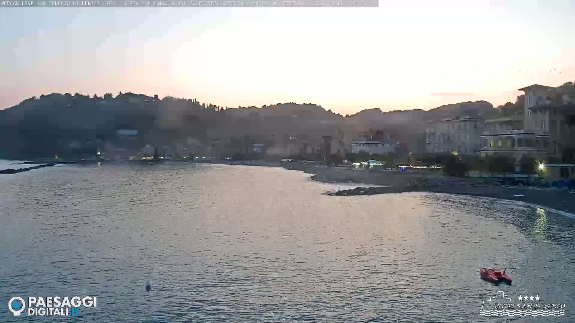 A calm body of water laps a dark-sand beach at sunset, with buildings and hills rising in the background.