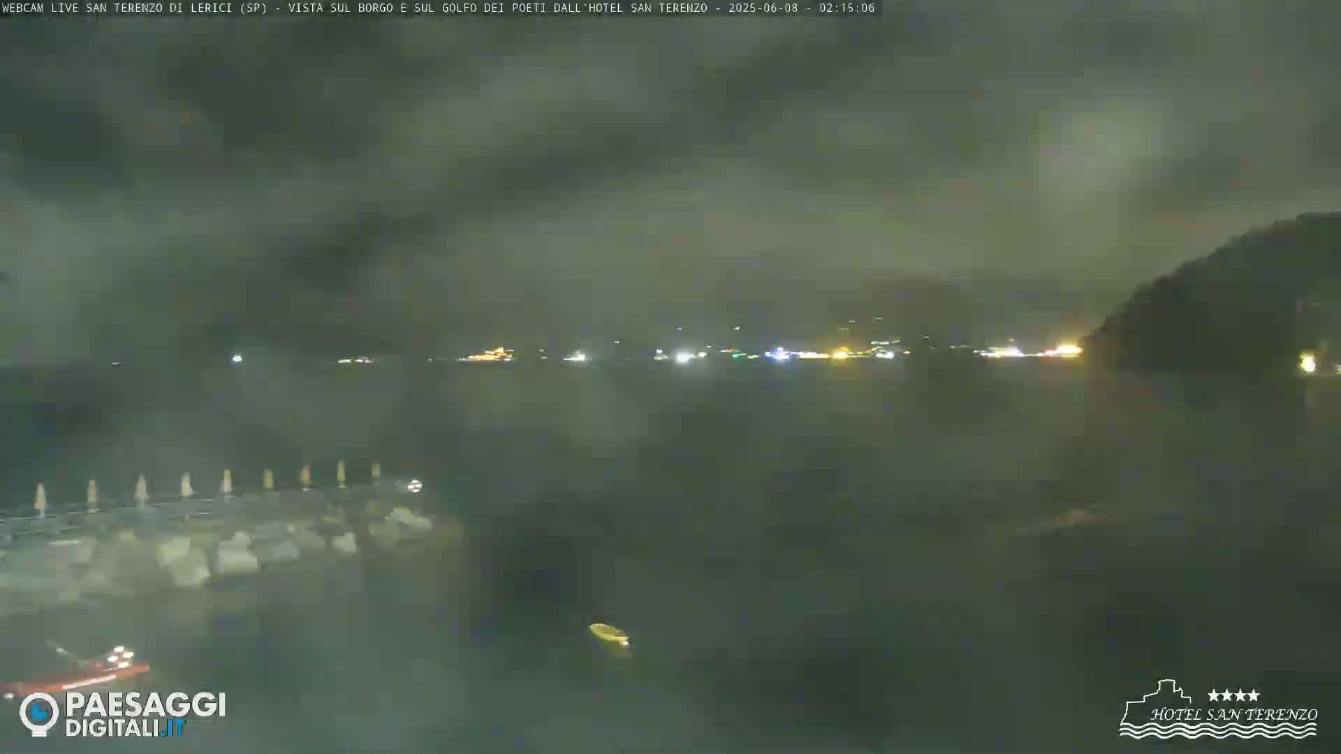 A nighttime coastal scene shows a dark body of water with faint lights across the horizon under a cloudy sky.