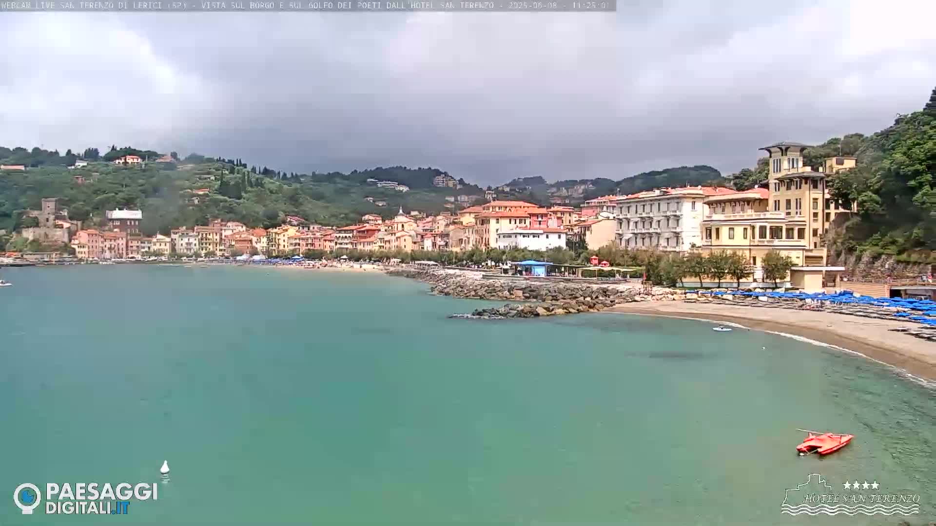 A coastal town with colorful buildings sits on a hillside overlooking a calm, teal bay with a sandy beach and scattered beach umbrellas under a cloudy sky.